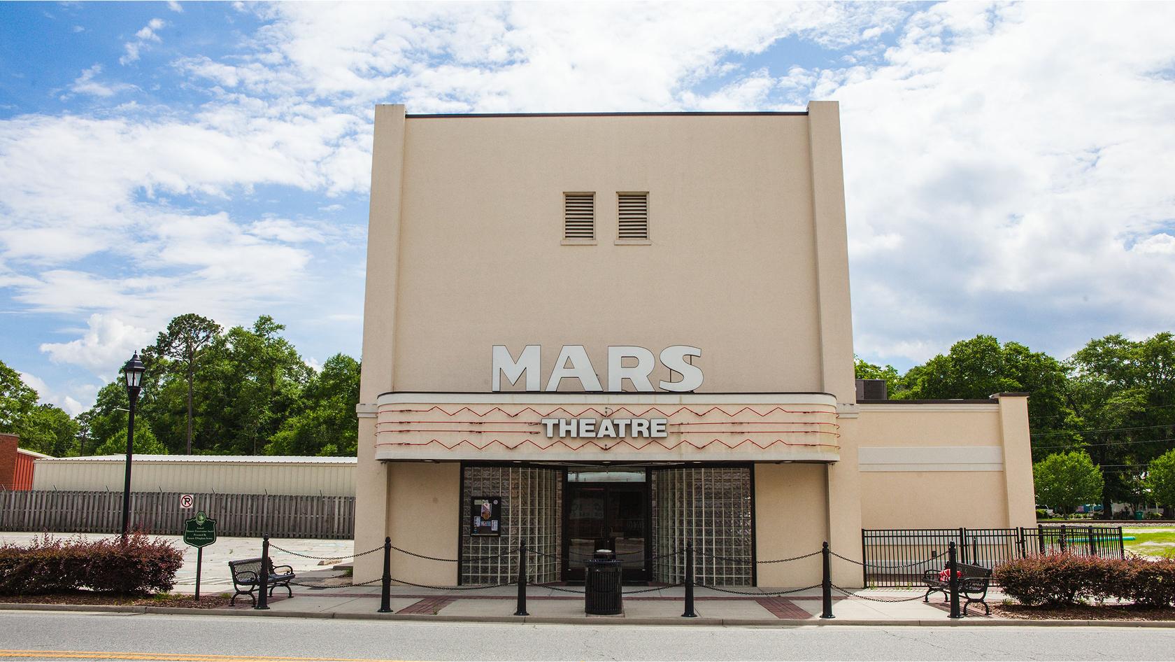 A beige, retro-style building with a marquee reading MARS THEATRE stands on a quiet street, surrounded by greenery and under a blue sky with scattered clouds.