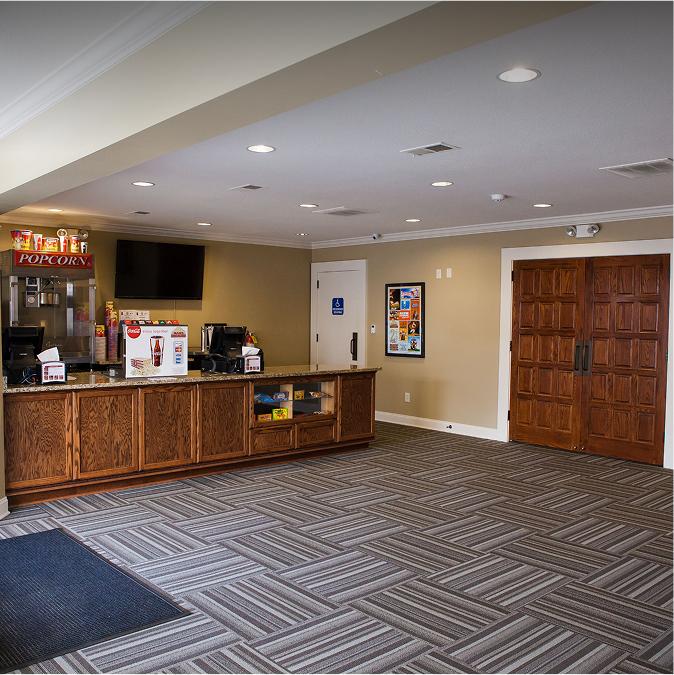 A lobby area with patterned carpet, wood double doors, and a concession counter displaying popcorn, snacks, and drinks. The walls are beige, and there’s a flat-screen TV above the counter.