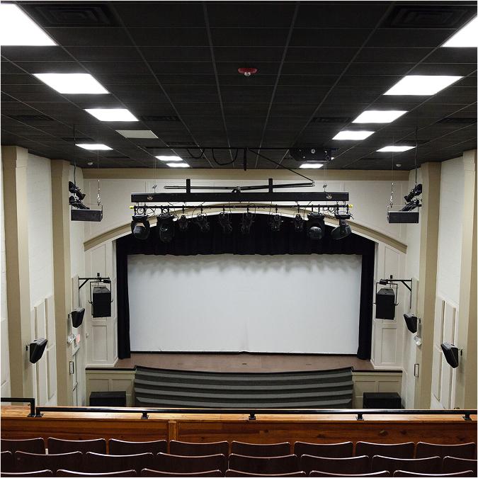 A view from the back of an empty auditorium showing rows of brown seats facing a stage with a white screen, black curtains, overhead lights, and sound equipment. The ceiling has a grid of lights and speakers.