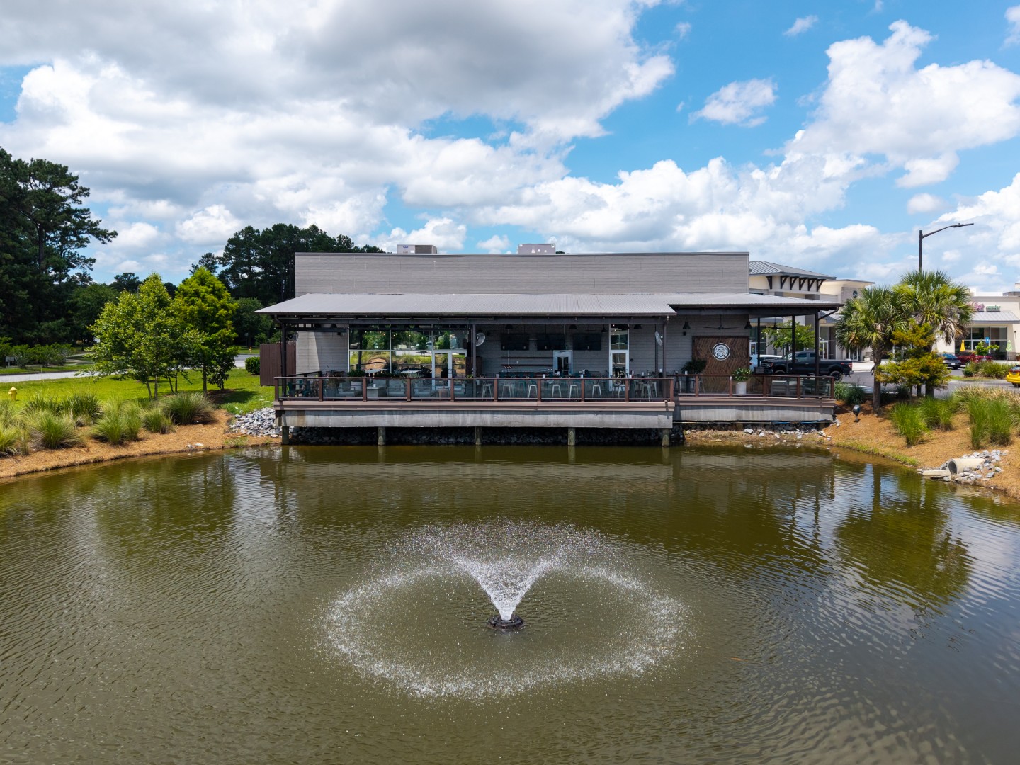 A restaurant with a covered outdoor patio overlooks a small pond with a water fountain, surrounded by greenery and trees under a partly cloudy blue sky.
