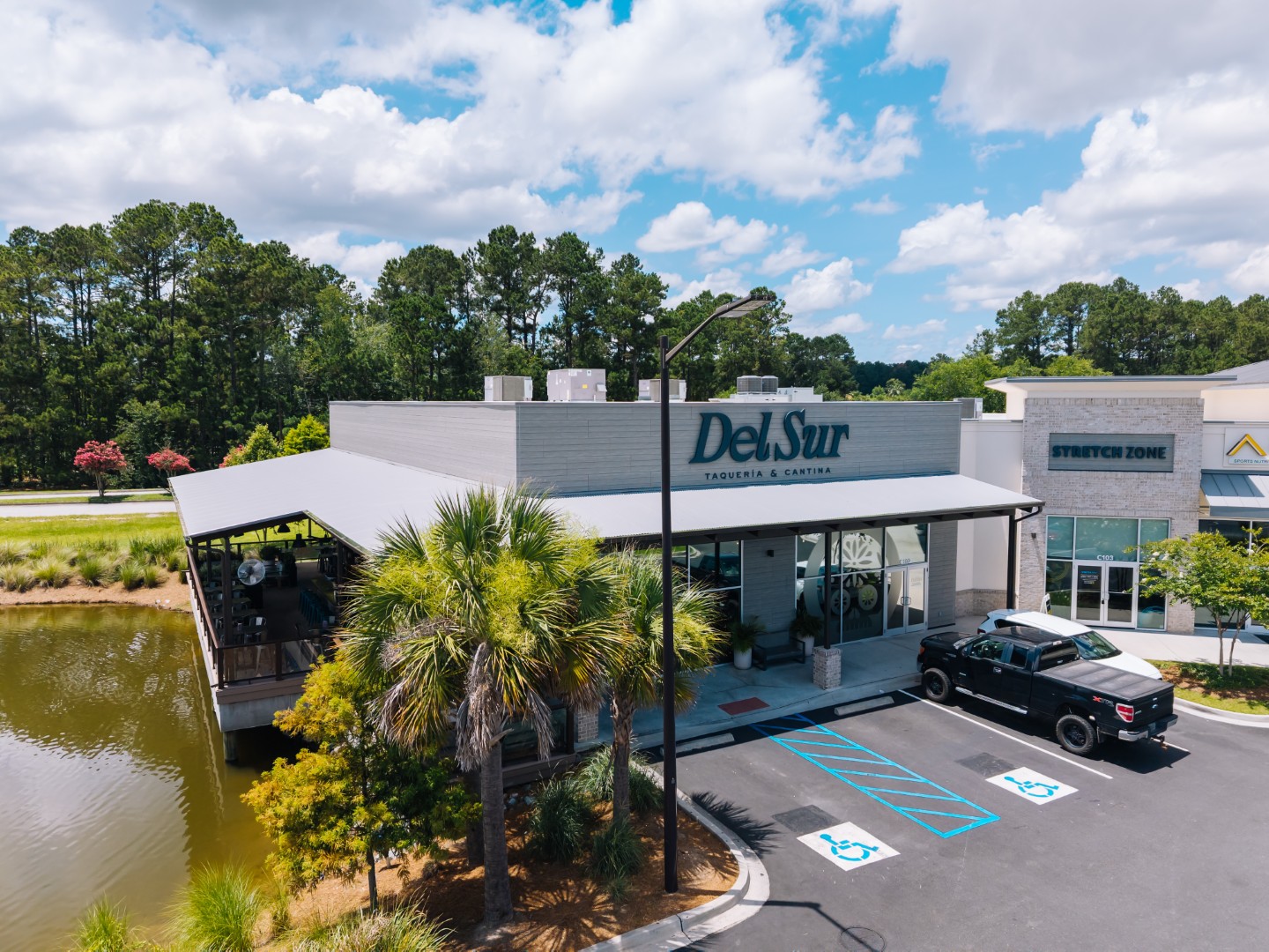 A modern restaurant called Del Sur Taqueria & Cantina is pictured with outdoor seating by a pond, palm trees, accessible parking spaces, and a black pickup truck parked in front under a partly cloudy sky.