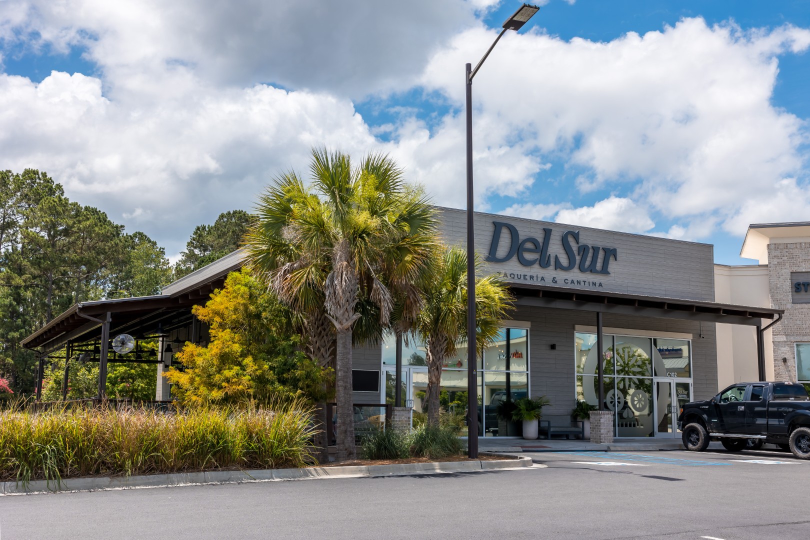 Exterior view of Del Sur Taqueria & Cantina restaurant, with palm trees and greenery in front, a modern facade, and a black truck parked nearby under a partly cloudy sky.
