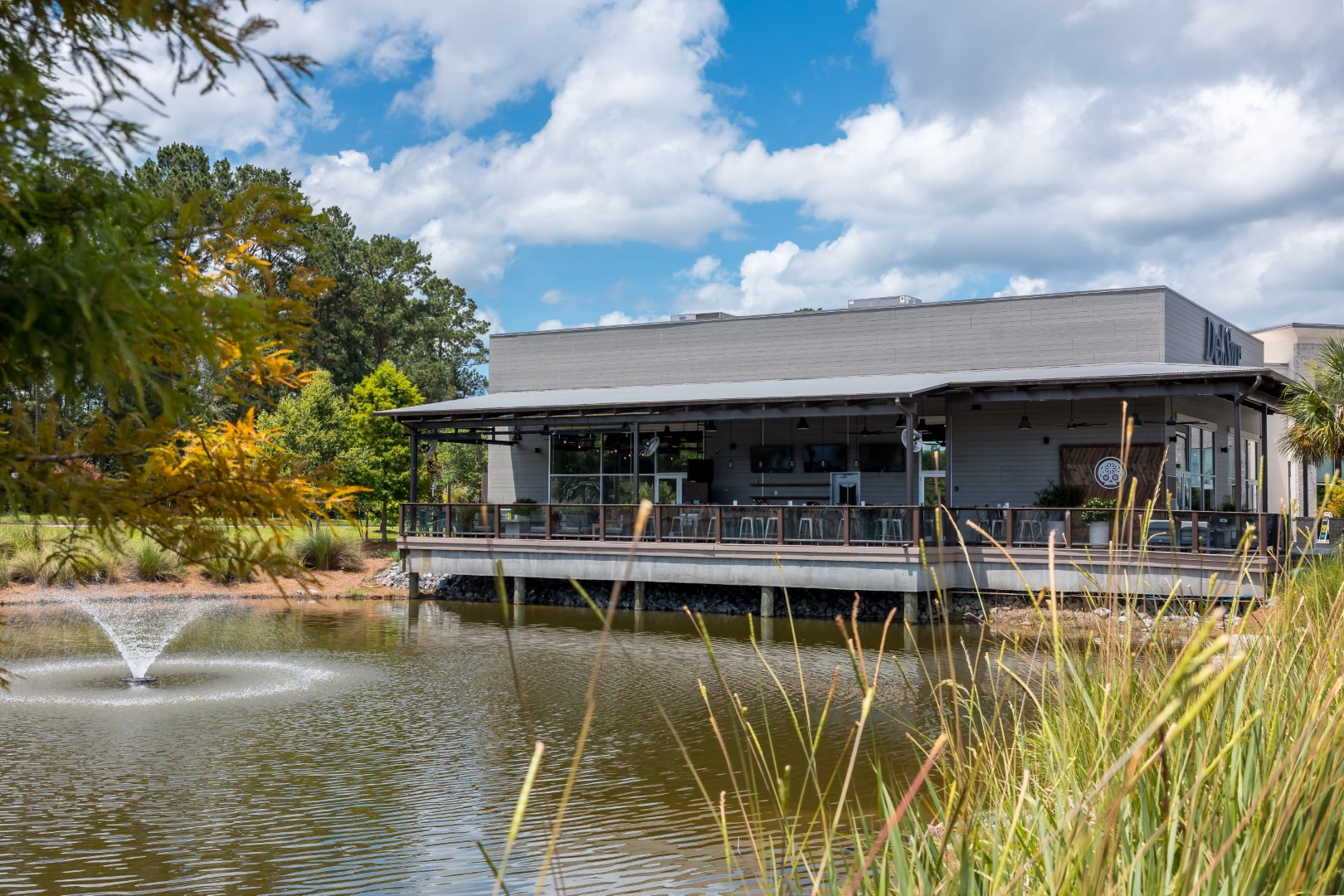 A building with a covered outdoor patio sits beside a pond with a small fountain, surrounded by grass and trees under a partly cloudy sky.