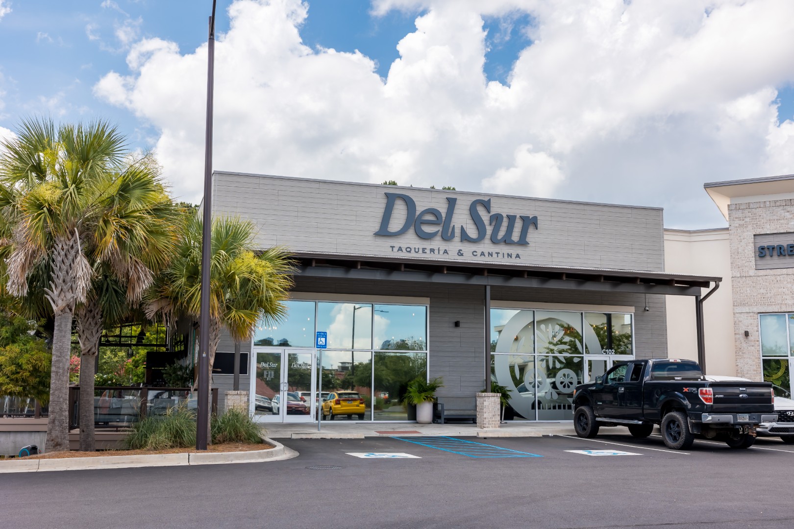 Exterior view of Del Sur Taqueria & Cantina, a restaurant with large glass windows, palm trees in front, and a black pickup truck parked in a nearby accessible parking space.