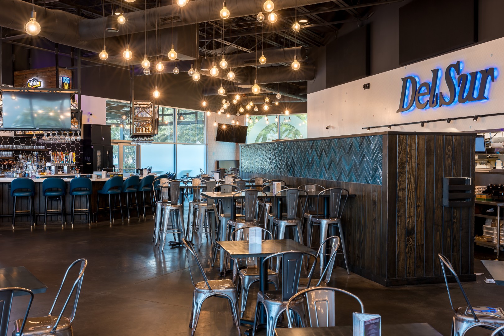 Spacious restaurant with metal tables and chairs, blue bar stools, exposed ceilings, hanging Edison bulbs, and a sign reading Del Sur on a wood-paneled wall. The bar area is visible in the background.
