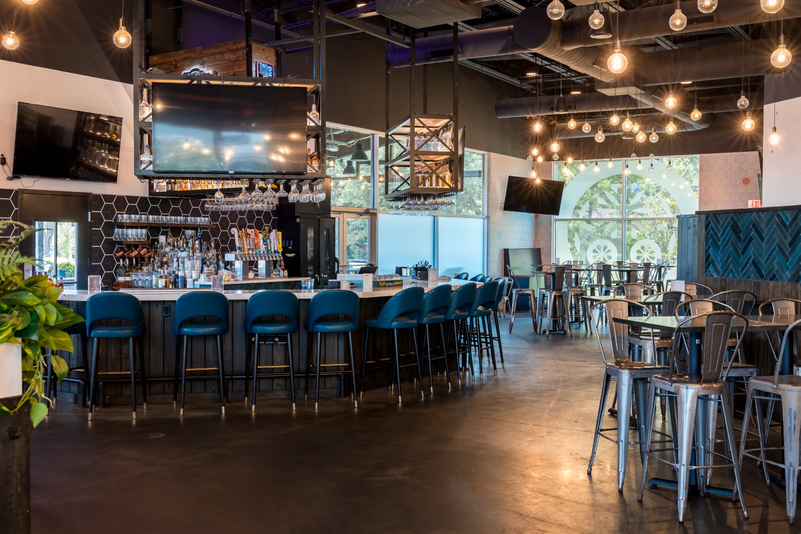 A bar with blue chairs around a curved counter, several TVs, hanging lights, and high tables with metal stools. Large windows let in natural light, creating an inviting and spacious atmosphere.