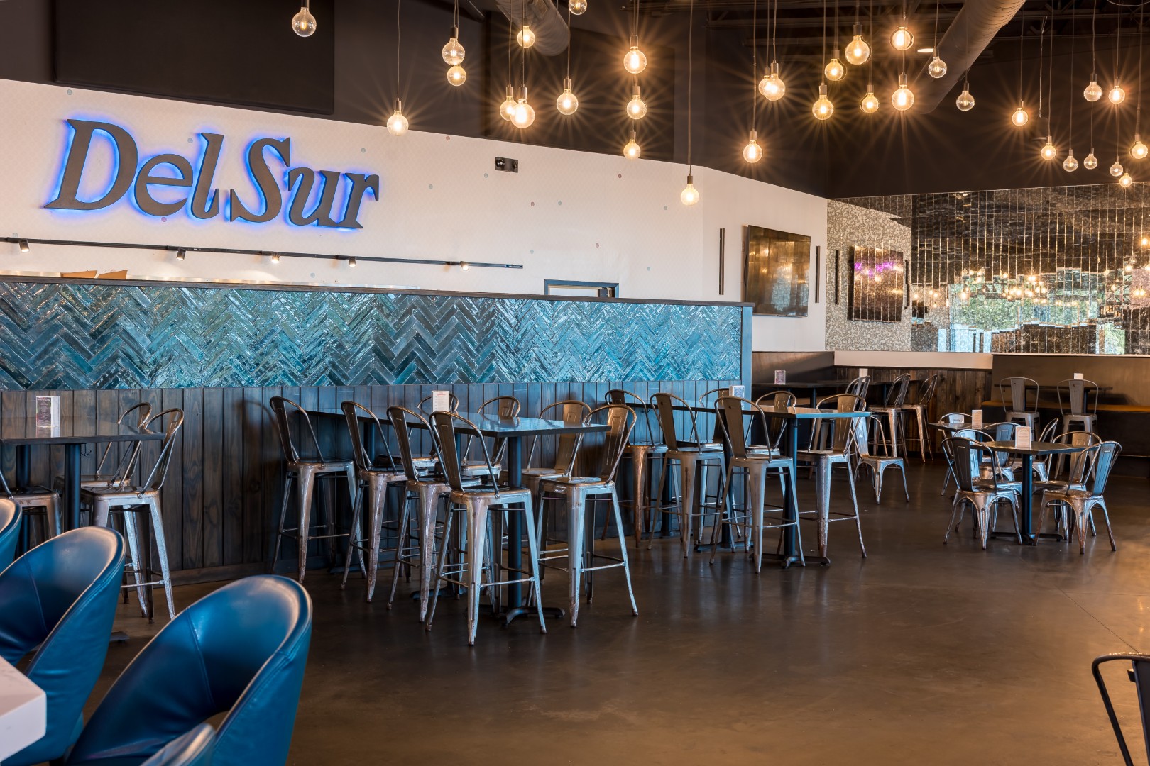 Restaurant interior with metal chairs and tables, blue accents, hanging light bulbs, a mirrored wall, and a neon Del Sur sign.