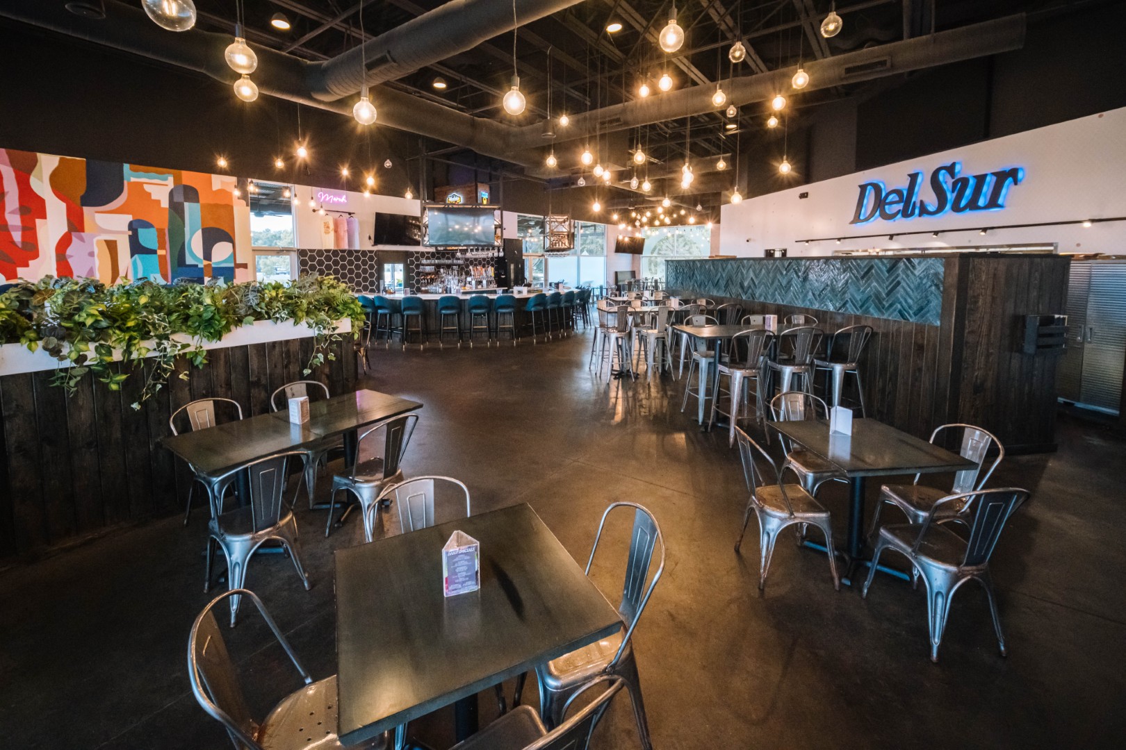 Restaurant interior with metal chairs and dark tables, hanging pendant lights, greenery along a divider, colorful wall art, a neon Del Sur sign, and a bar area with high stools in the background.