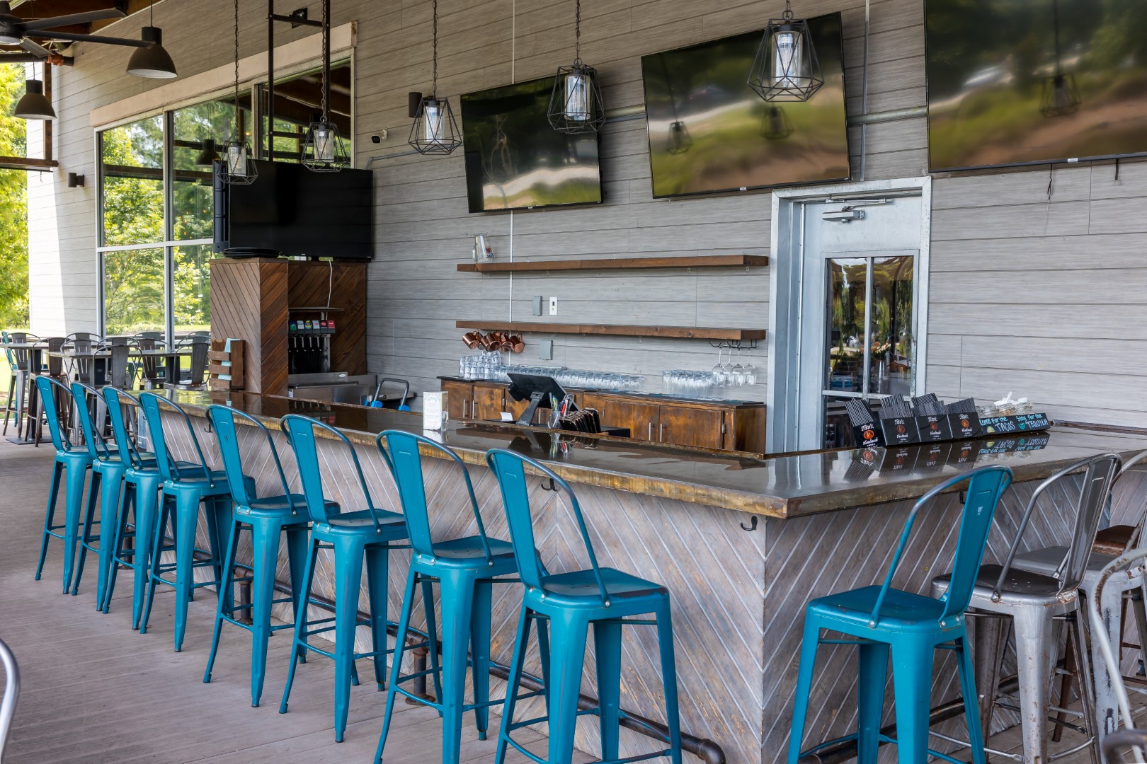 Outdoor bar with a row of blue metal stools along a wooden counter, glassware on shelves, several flat-screen TVs on the wall, and bright natural light coming from large windows.