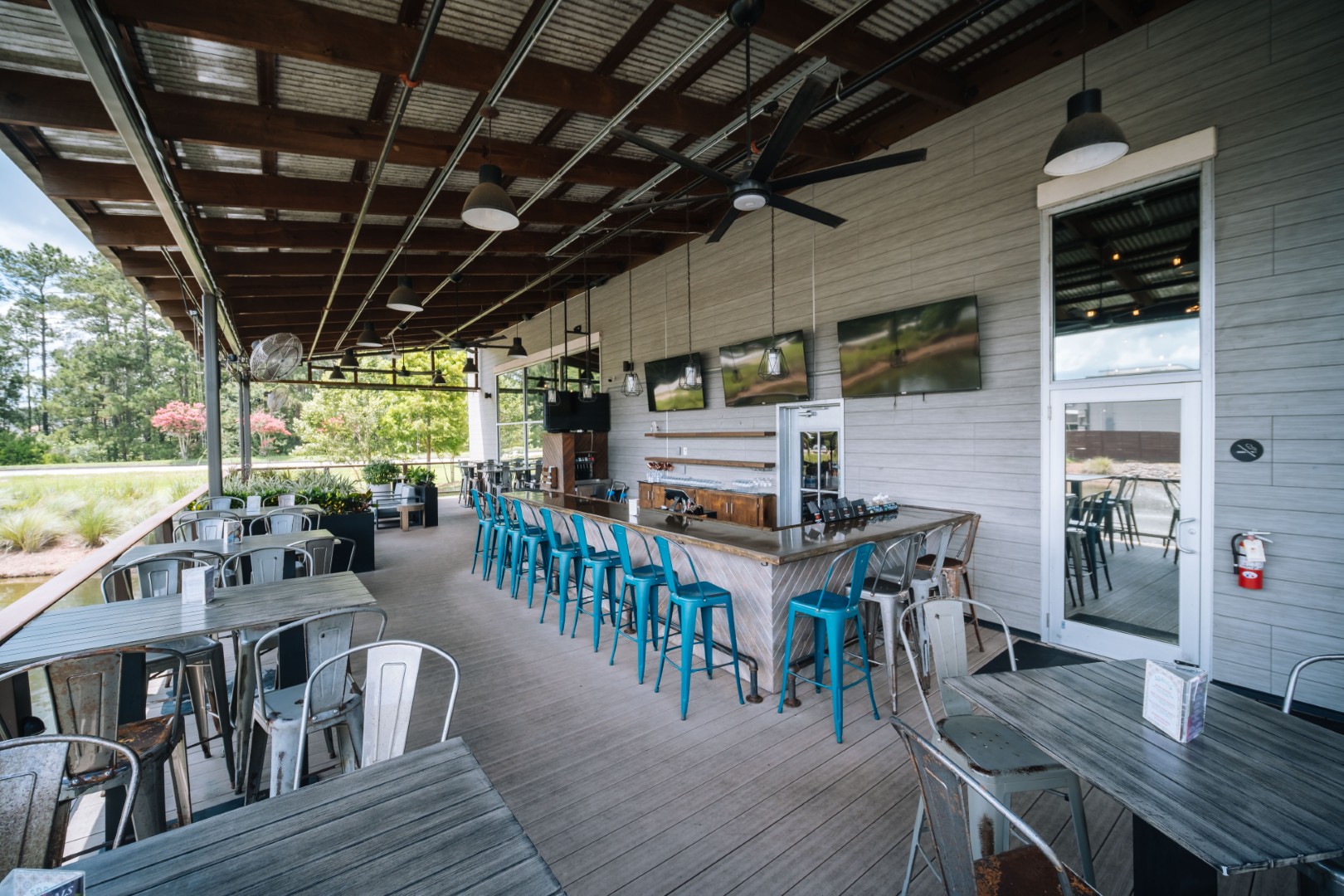 An outdoor bar and dining area with metal tables and chairs, a long bar with blue stools, ceiling fans, and several TVs mounted on the wall. The space is covered with a wooden roof and overlooks a green landscape.