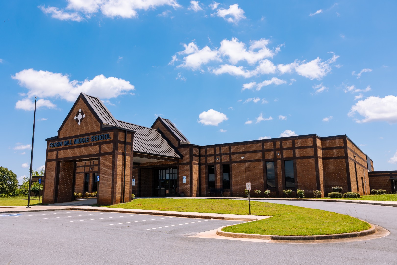 A brick school building labeled Feagin Hill Middle School sits under a bright blue sky with scattered clouds. The parking lot in front is empty, and the landscaping is neatly maintained.