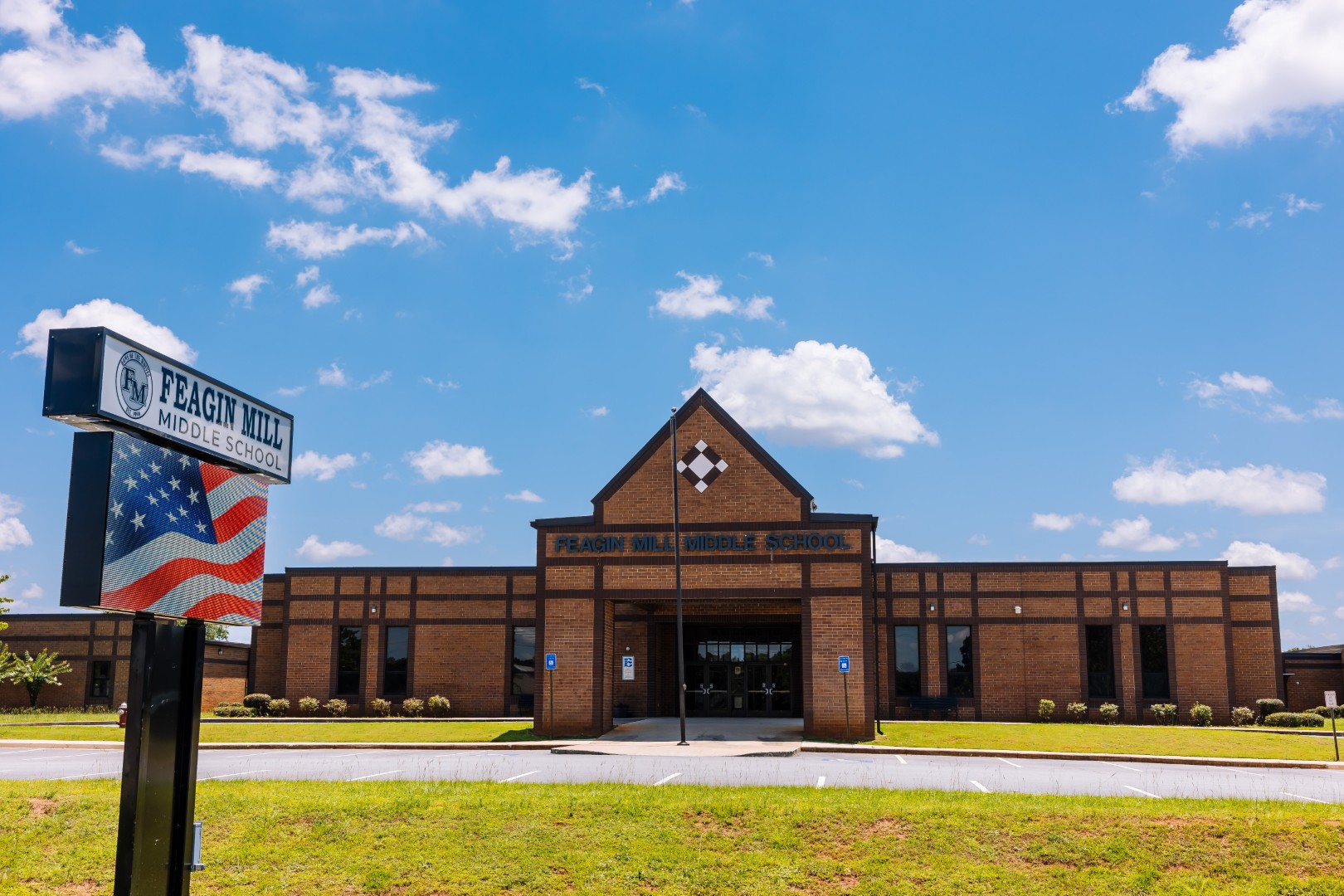 A brick building with a sign reading Feagin Mill Middle School stands under a blue sky with scattered clouds. An American flag is shown on the sign in front of the school.