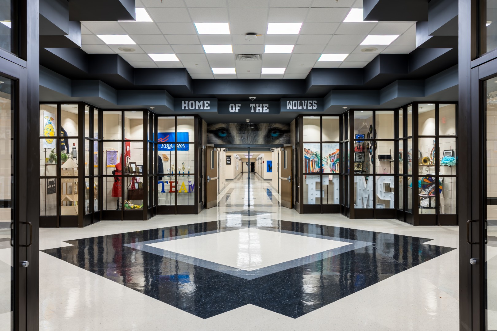 A bright school hallway with display cases of student artwork and trophies, a geometric black and white floor, and a sign above the hallway reading Home of the Wolves with large wolf eyes depicted above it.