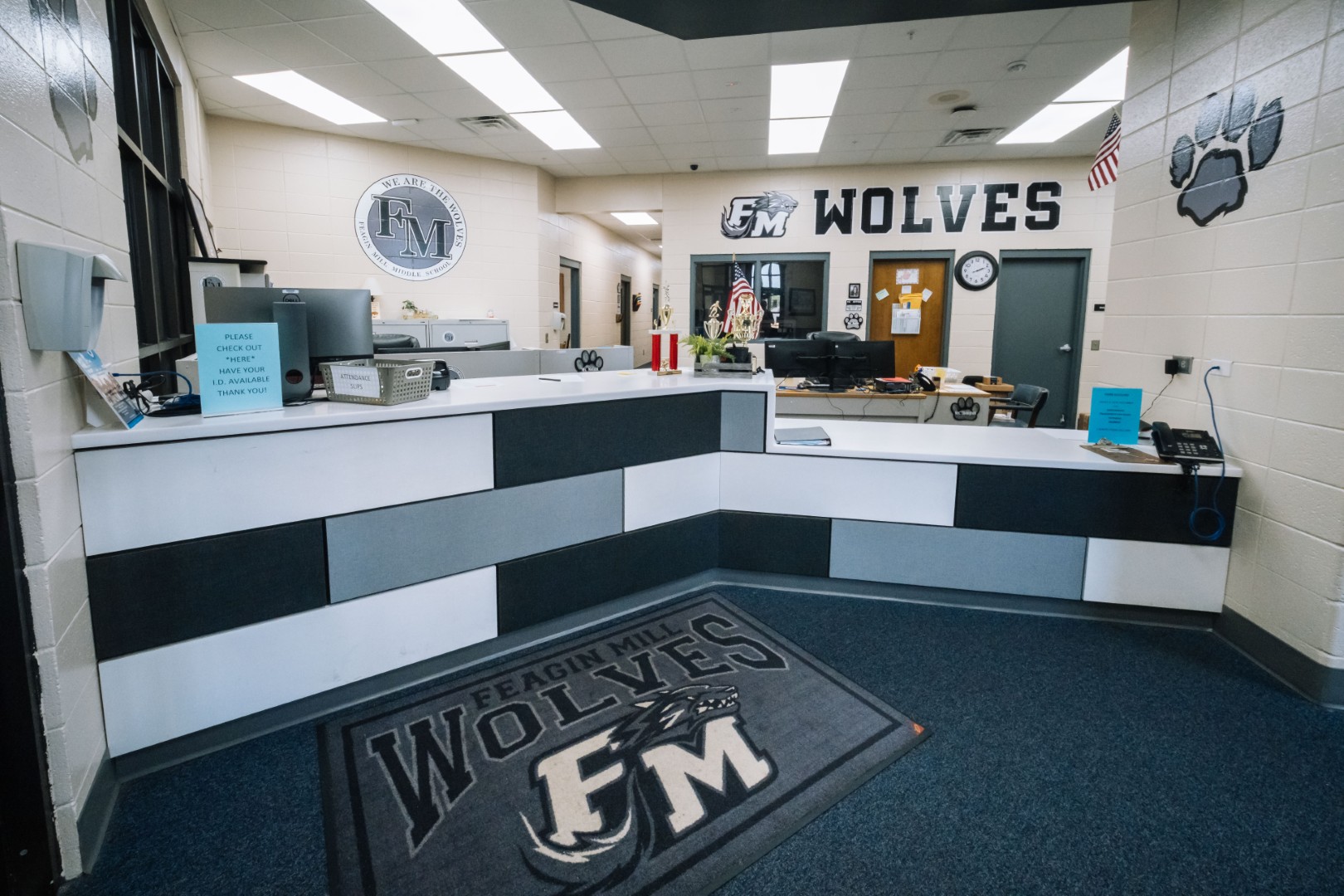 A school office reception area with a black, white, and gray front desk, “FM Wolves” logos on the wall and floor, and office supplies, signs, and papers on the counter.