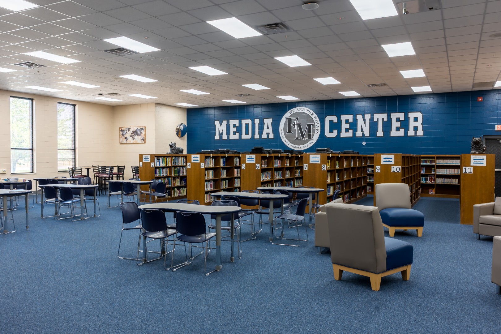 A spacious library with blue carpet, bookshelves, tables, chairs, and a wall labeled “MEDIA CENTER.” Large windows let in natural light and the room appears organized and welcoming.