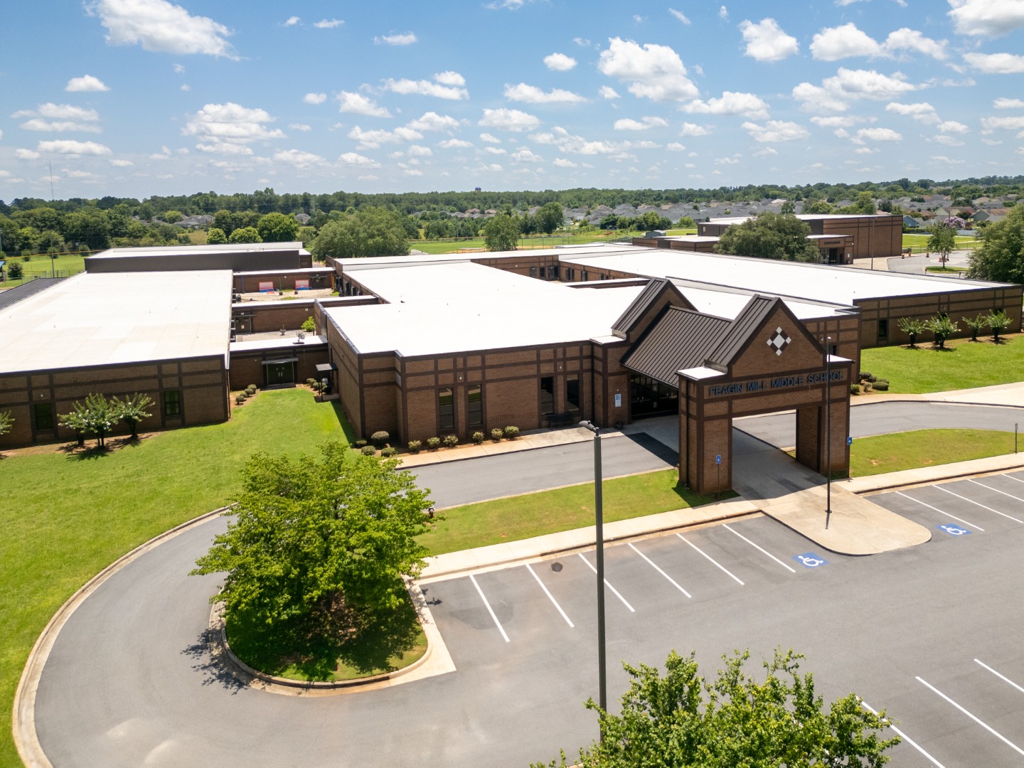 Aerial view of a large, modern school building with a brick exterior, surrounded by a parking lot, green lawns, and trees under a blue sky with scattered clouds.