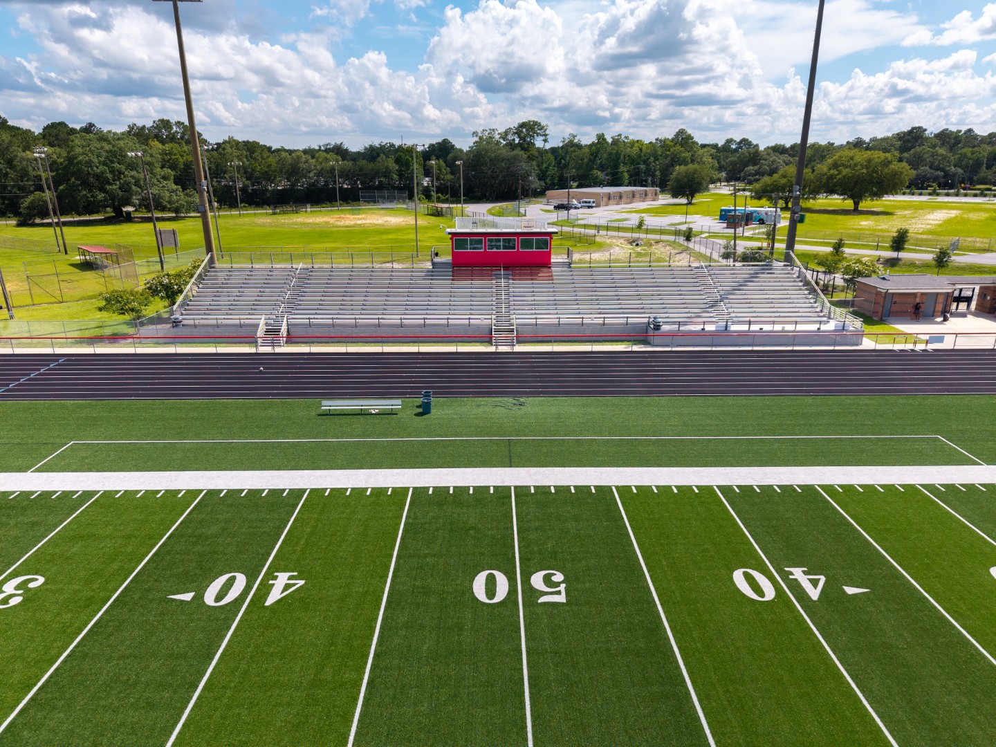 A football field with white yard lines and numbers, empty metal bleachers, a red press box, and light poles under a partly cloudy sky, surrounded by green grass and trees.