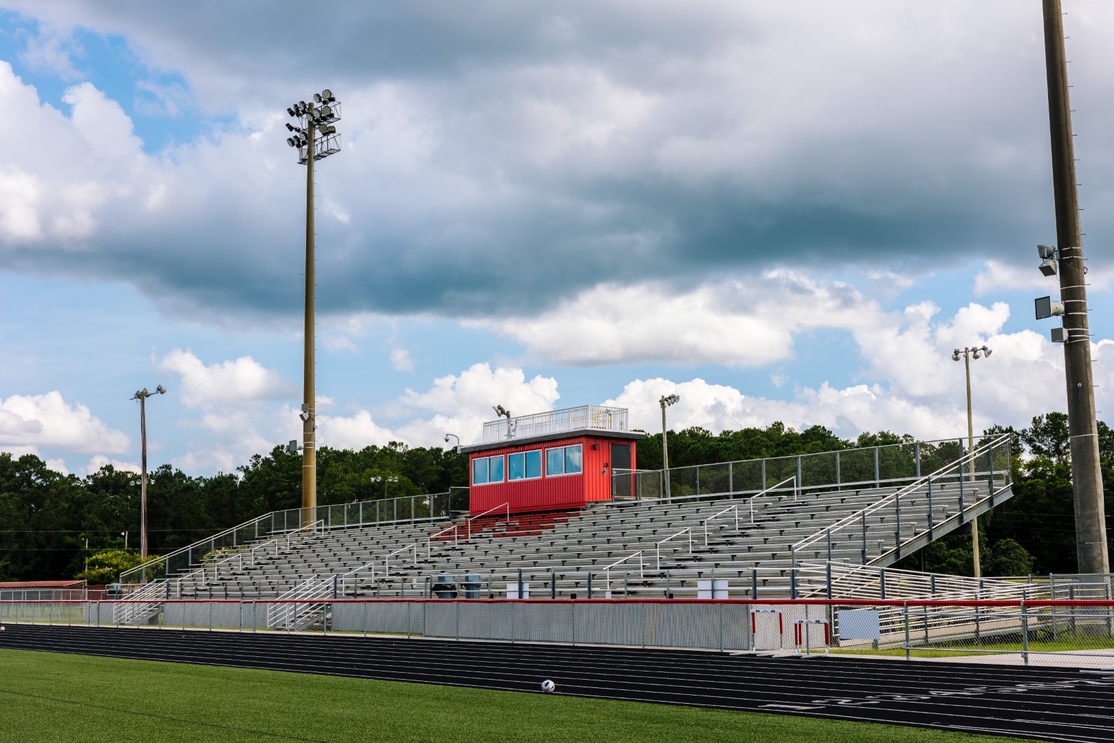 Empty outdoor bleachers with a red press box overlook a track and football field under a partly cloudy sky. Tall light poles border the stands and trees line the background.
