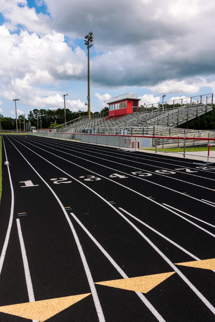 A black running track with numbered lanes stretches toward empty bleachers and a red press box under a partly cloudy sky at an outdoor stadium.