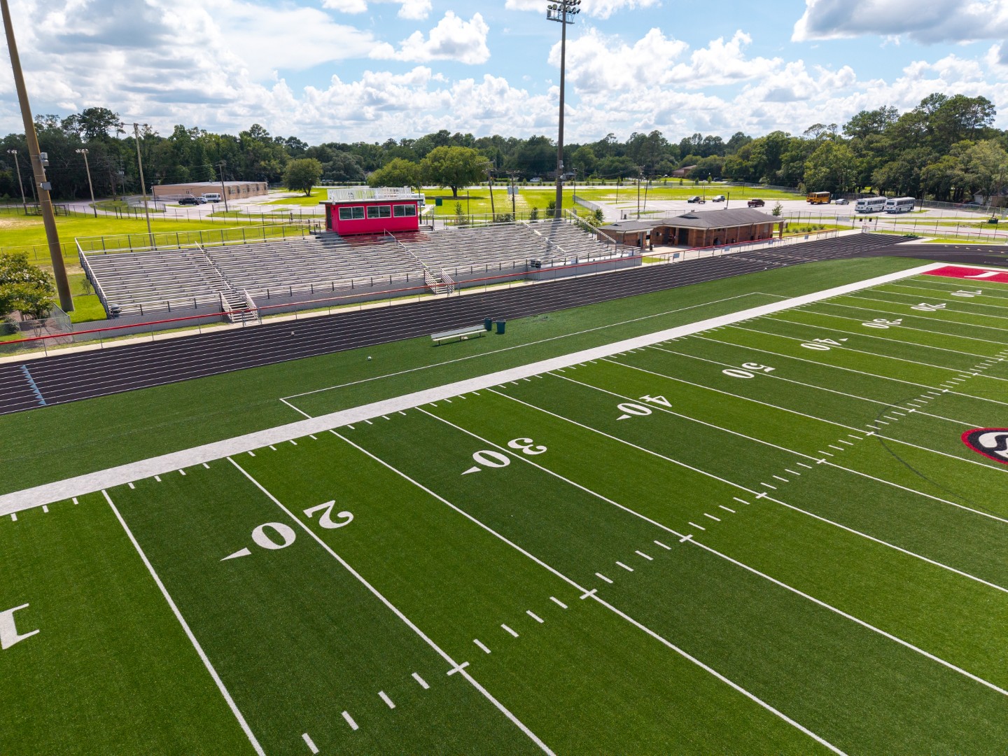 A wide view of an empty football field with white yard lines, green artificial turf, and bleachers in the background under a partly cloudy sky. A red press box sits above the bleachers, and trees surround the area.