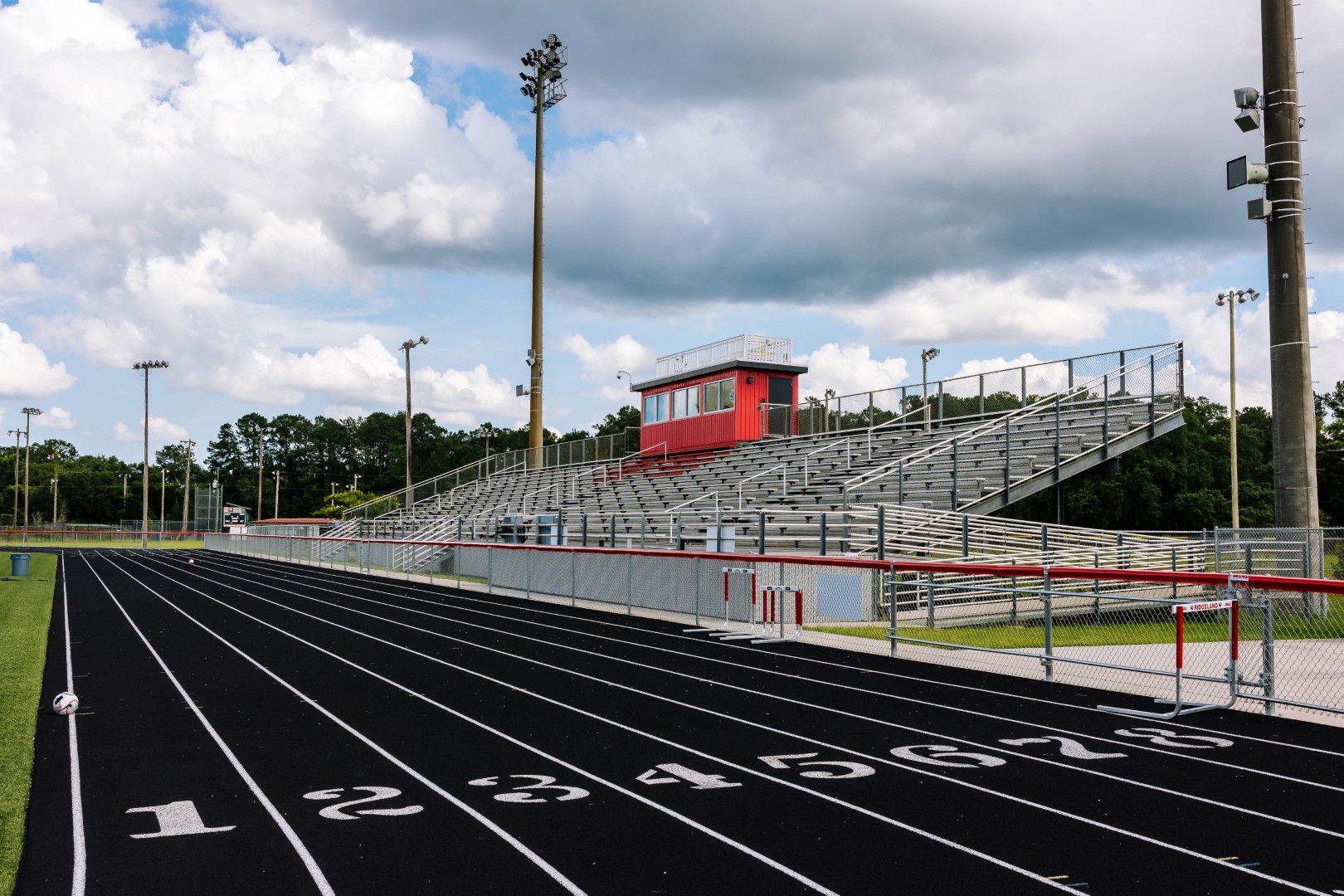 A black running track with white lane numbers curves past a red and white bleacher stand under cloudy skies at an empty outdoor stadium.