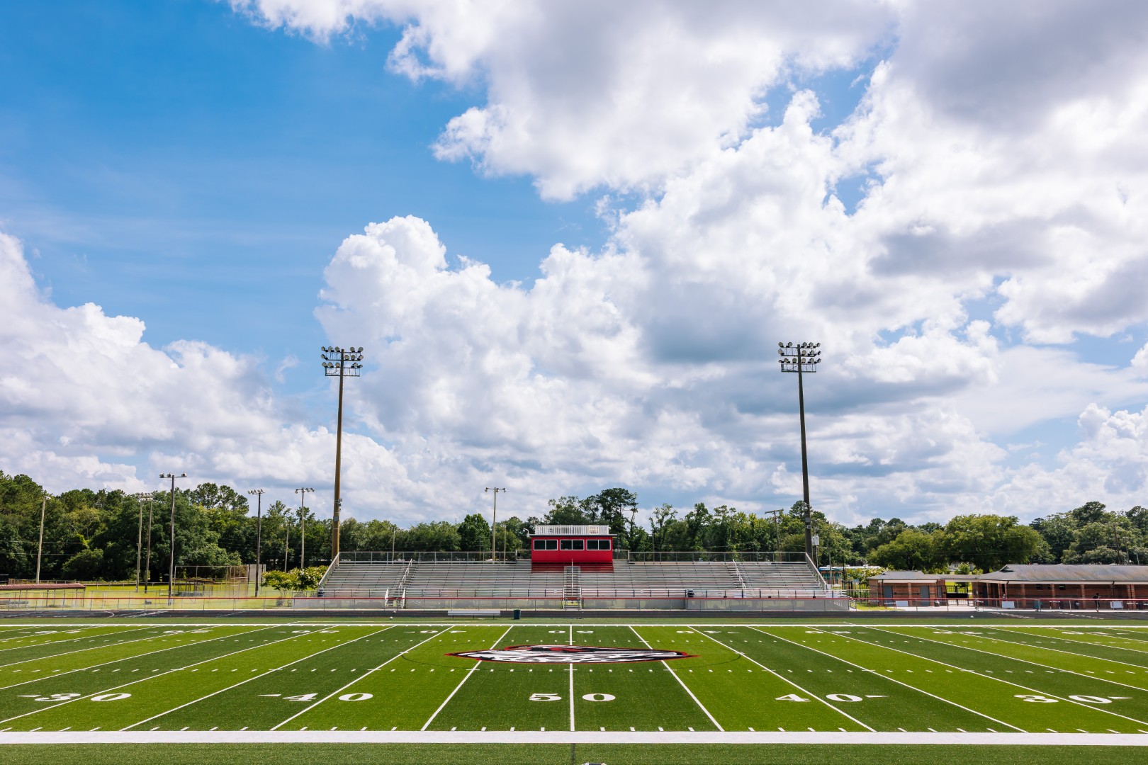 A wide view of an empty football field with white yard lines, a red and white press box, and bleachers under a partly cloudy sky. Tall light poles surround the field, and trees are visible in the background.