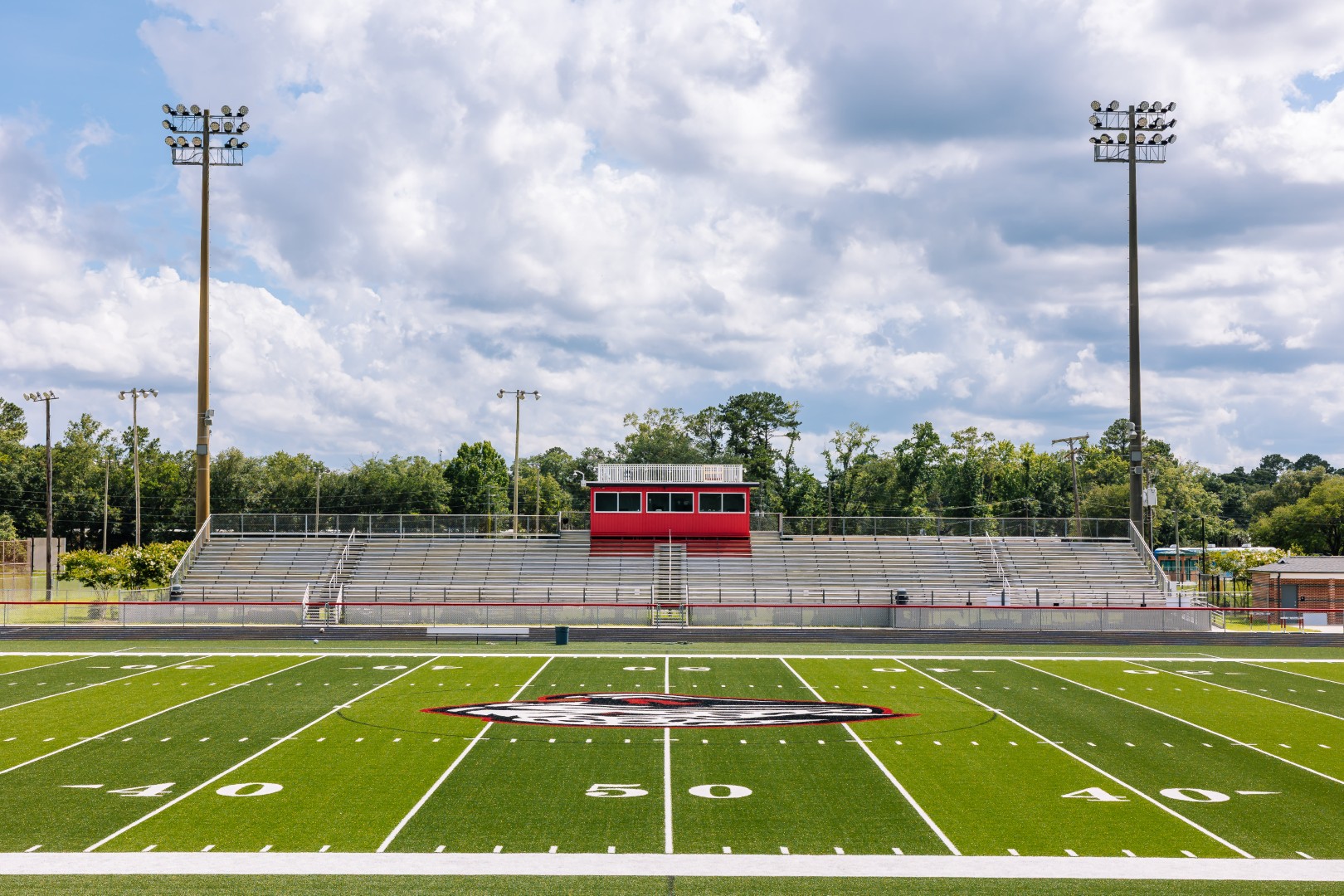 A high school football field with bright green turf, white yard lines, and a red and black logo at the center. Empty metal bleachers and a red press box are in the background under a partly cloudy sky.