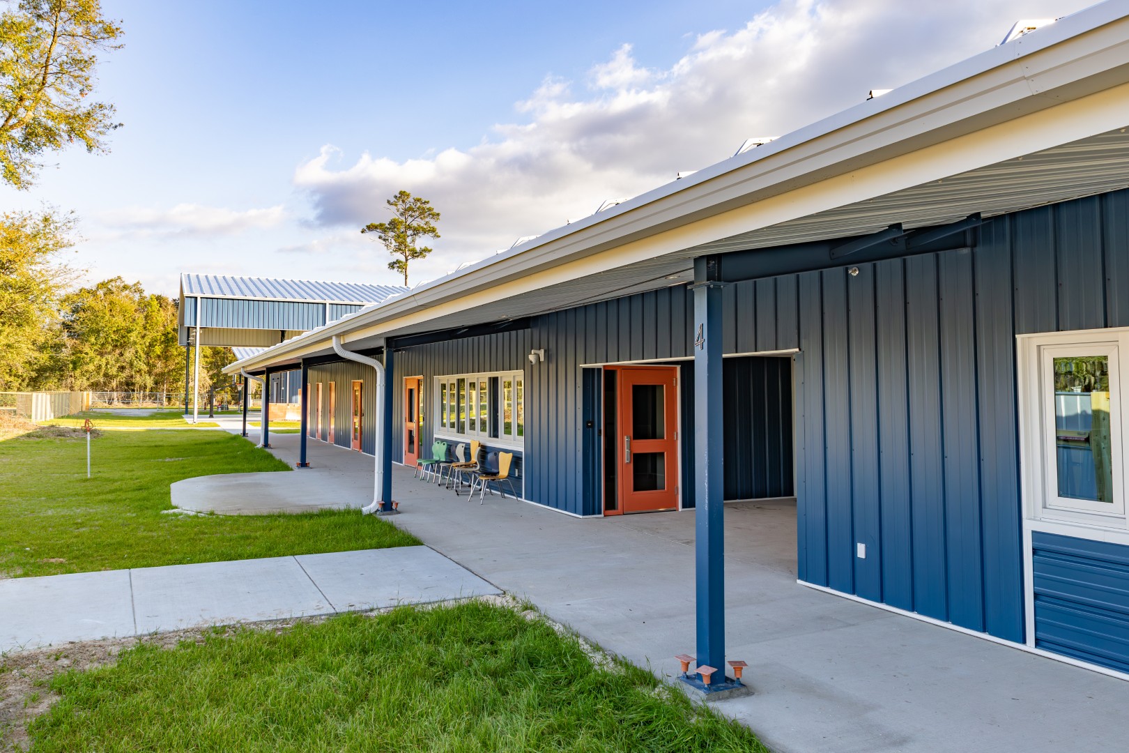 A single-story building with blue metal siding, orange doors, and white window frames. There are chairs and tables outside along a covered walkway, with grass and trees in the background under a partly cloudy sky.