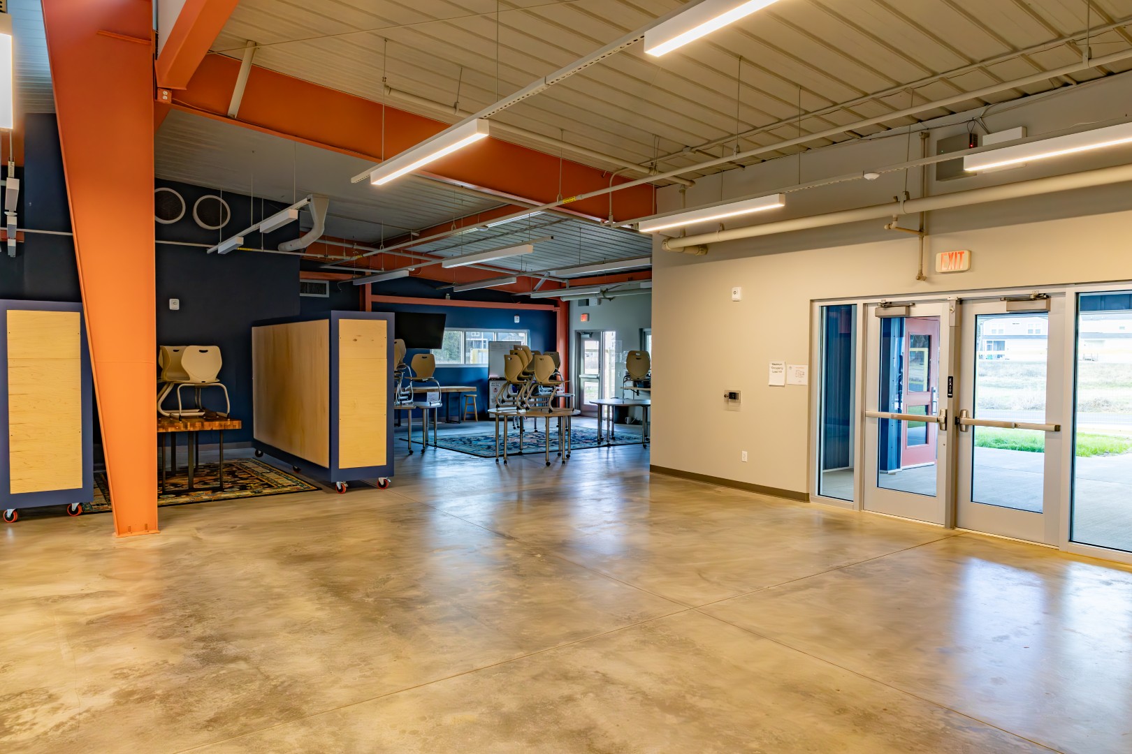 A spacious room with concrete floors, orange beams, stacked chairs on tables, large cabinets, and glass doors allowing natural light to enter. The space appears to be a classroom or multipurpose area.