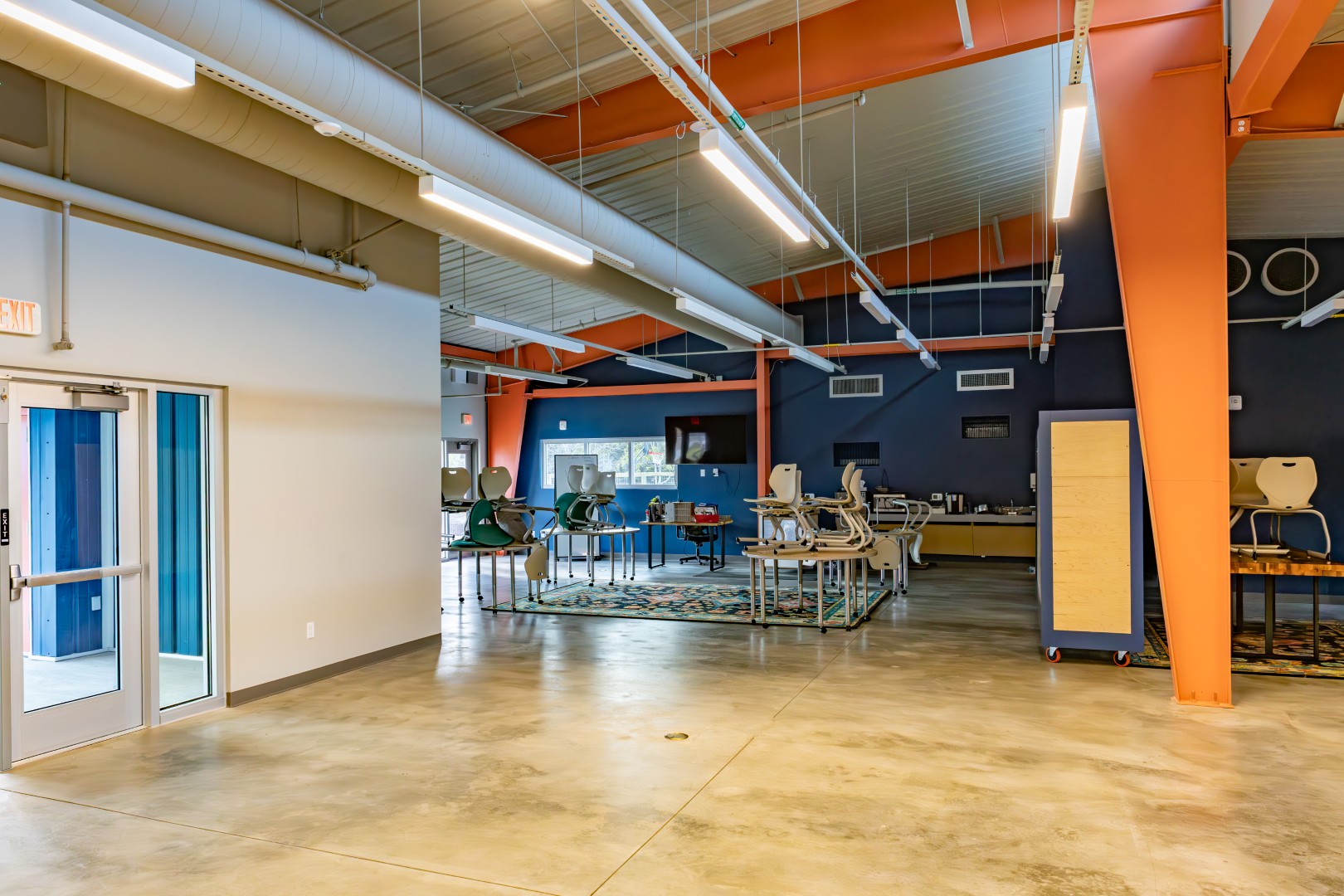 A spacious room with polished concrete floors, orange beams, blue and gray walls, tables and stacked chairs, large windows, and overhead lighting, resembling a collaborative workspace or classroom.