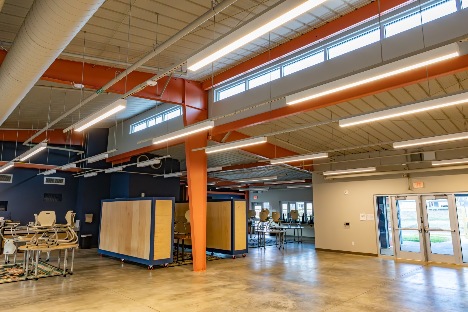 A spacious room with high ceilings, large windows, exposed beams, and overhead lighting. There are tables and chairs, glass doors leading outside, and a mix of blue, orange, and beige walls.