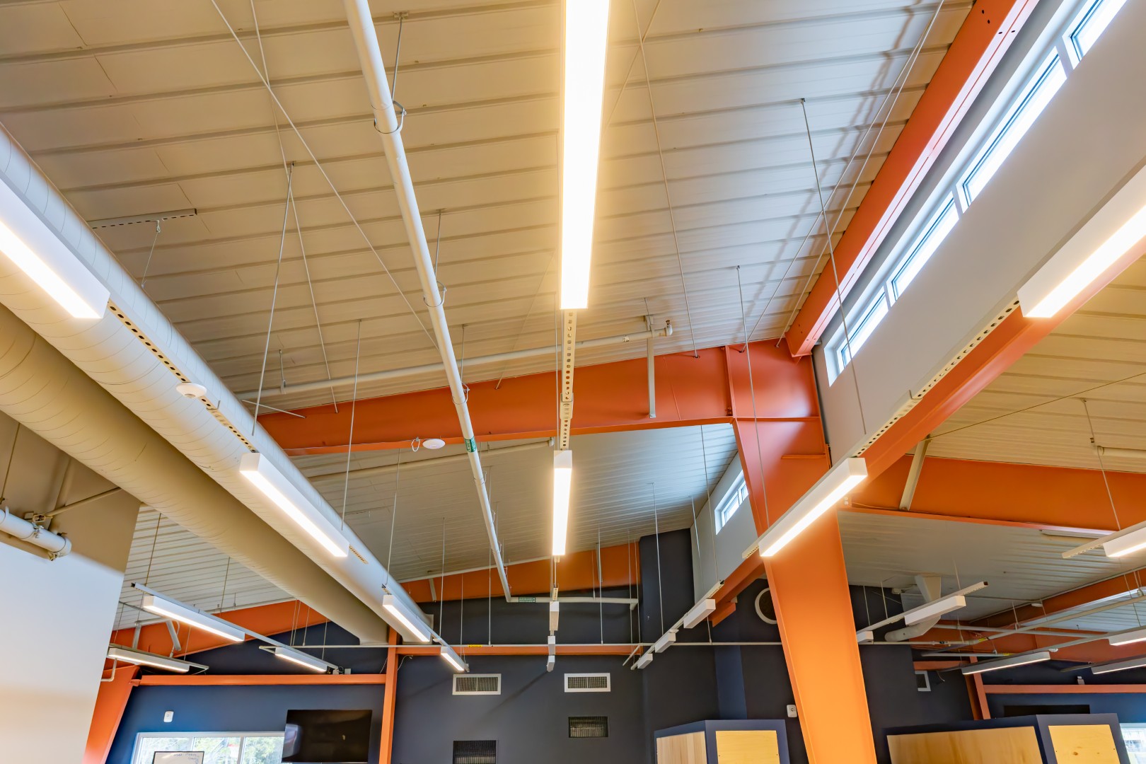 Ceiling of a building with exposed pipes, beams, and ductwork, featuring bright linear LED lights and clerestory windows letting in natural light.