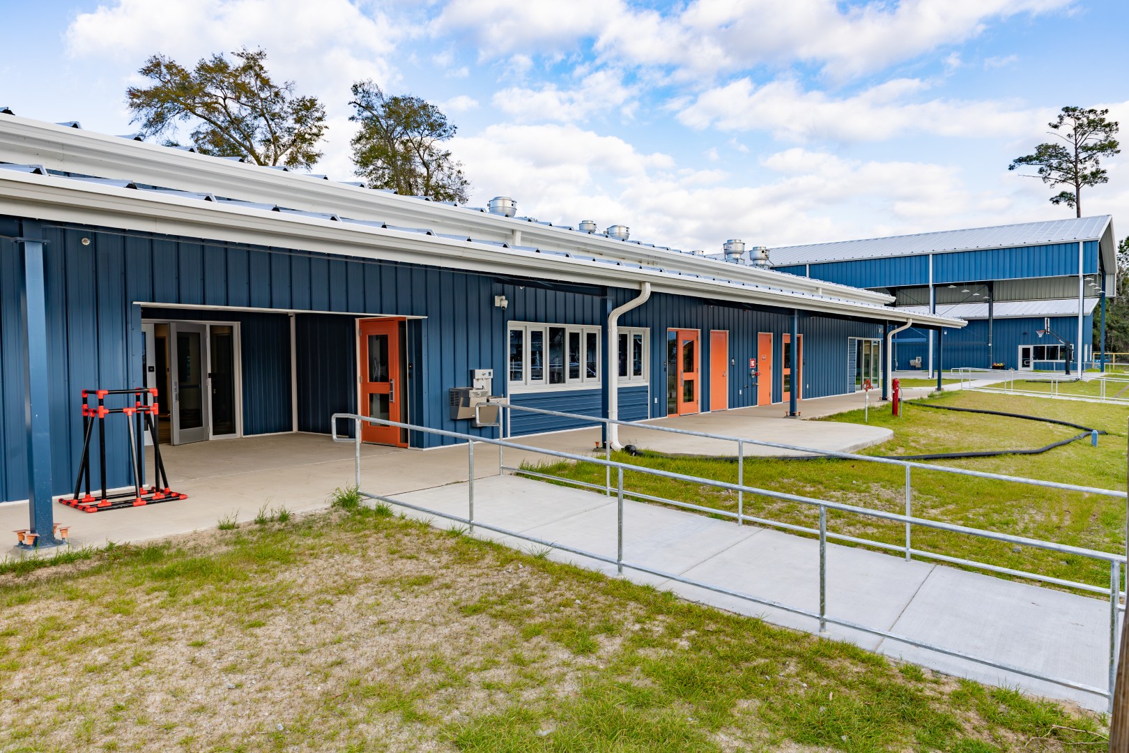 A blue metal building with orange-trimmed doors, large windows, and accessible ramps, surrounded by grass and trees under a partly cloudy sky.