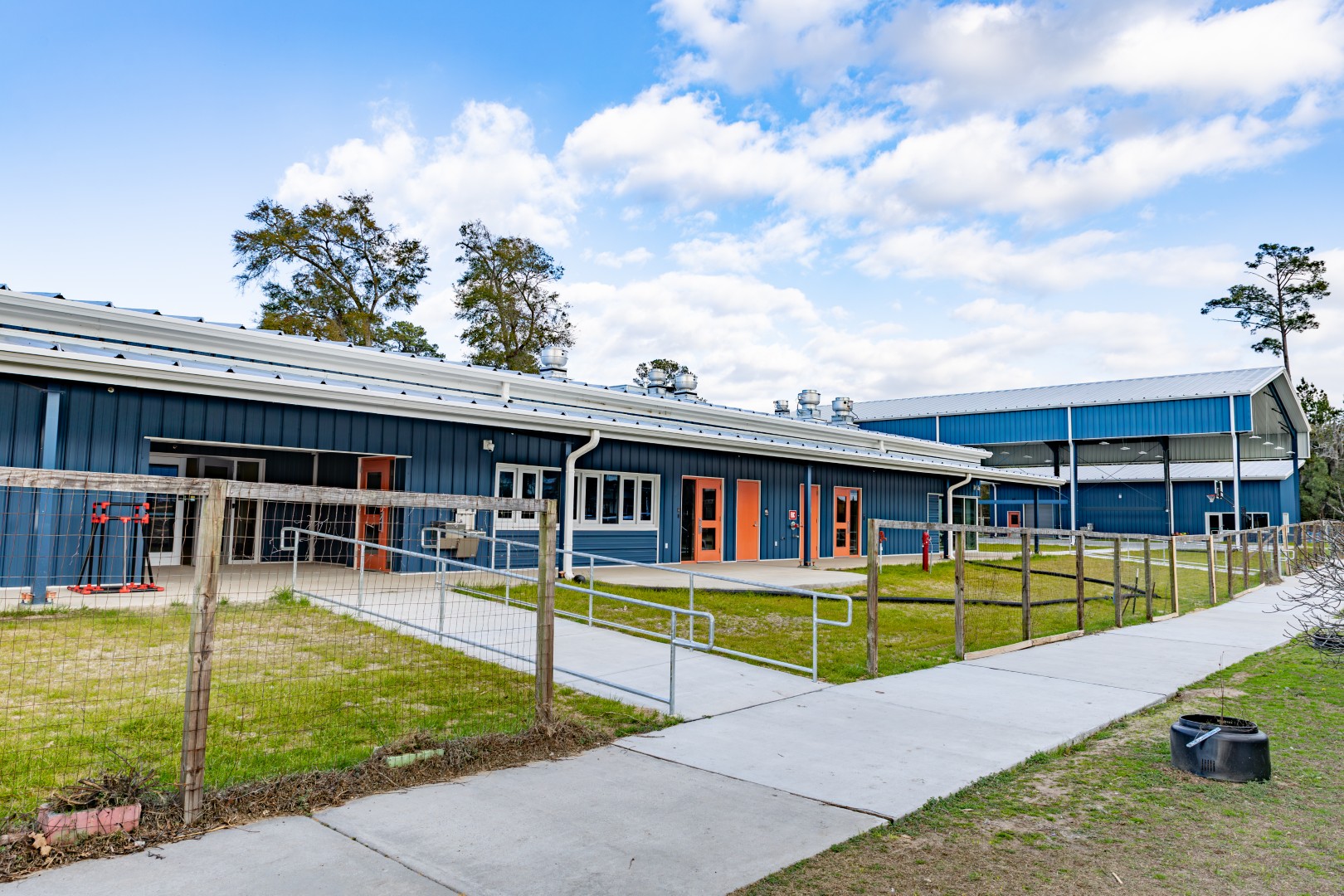 A blue building with orange doors, fenced grassy areas, and a sidewalk under a partly cloudy sky. Tall trees surround the building, and outdoor equipment is visible near the entrance.