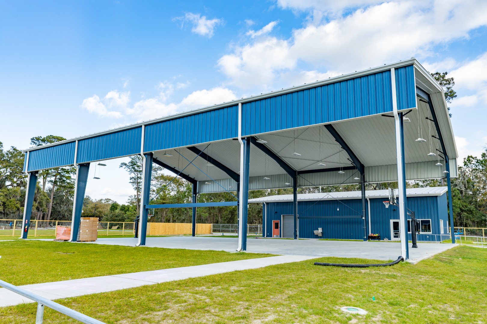 A large open-sided blue metal pavilion with a high roof stands on a grassy area under a partly cloudy sky, surrounded by trees and fenced areas. A paved walkway leads to the structure.