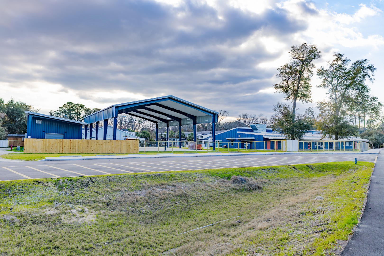 A large blue metal pavilion next to a school building with blue trim, an empty parking lot, and trees in the background under a partly cloudy sky.
