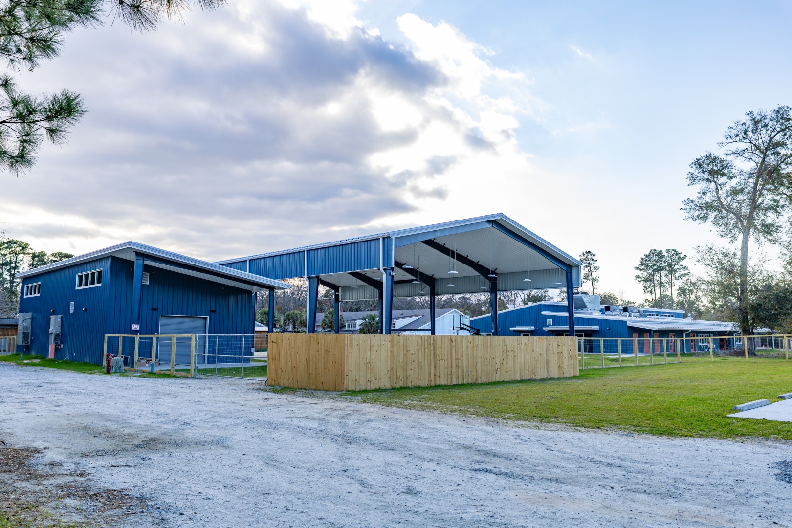 A blue barn with a large covered outdoor area and fenced enclosures, surrounded by grass and trees under a partly cloudy sky.