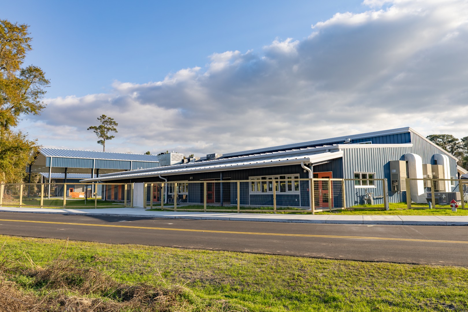 A single-story school building with large windows and metal siding, surrounded by a fence, sits under a partly cloudy sky with grass and a paved road in the foreground.