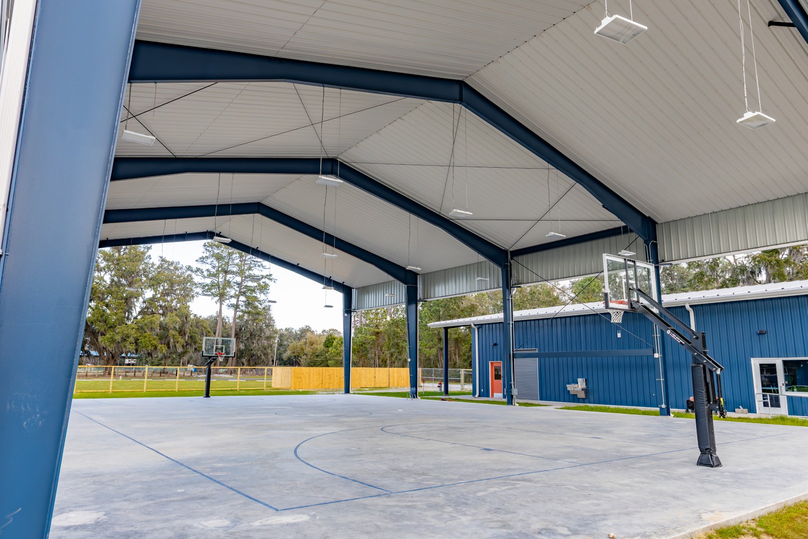Covered outdoor basketball court with multiple hoops, blue structural beams, and a metal roof. The area is open on the sides, with a blue building and green grass visible in the background.