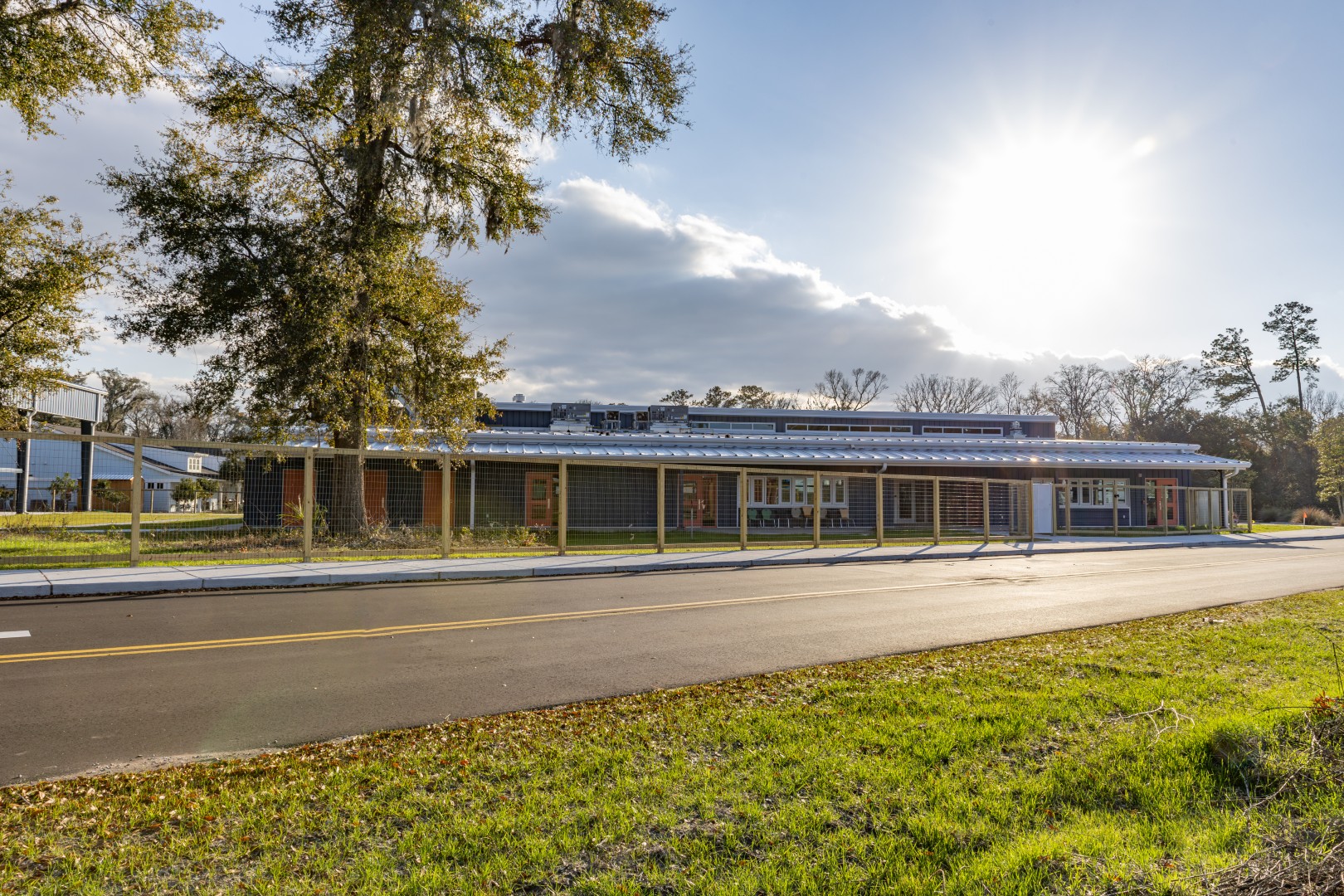A single-story building sits behind a covered walkway on a quiet road, with green grass and trees in the foreground and the sun shining brightly in a partly cloudy sky.