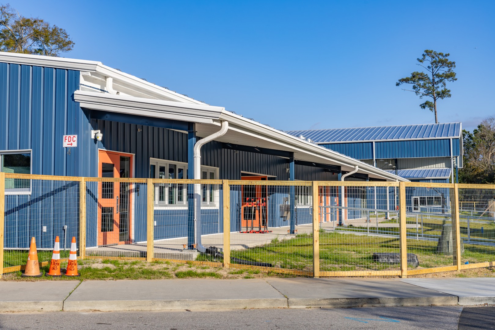 A blue building with orange doors and white trim is behind a yellow temporary construction fence. Three orange traffic cones are placed near the sidewalk in front.