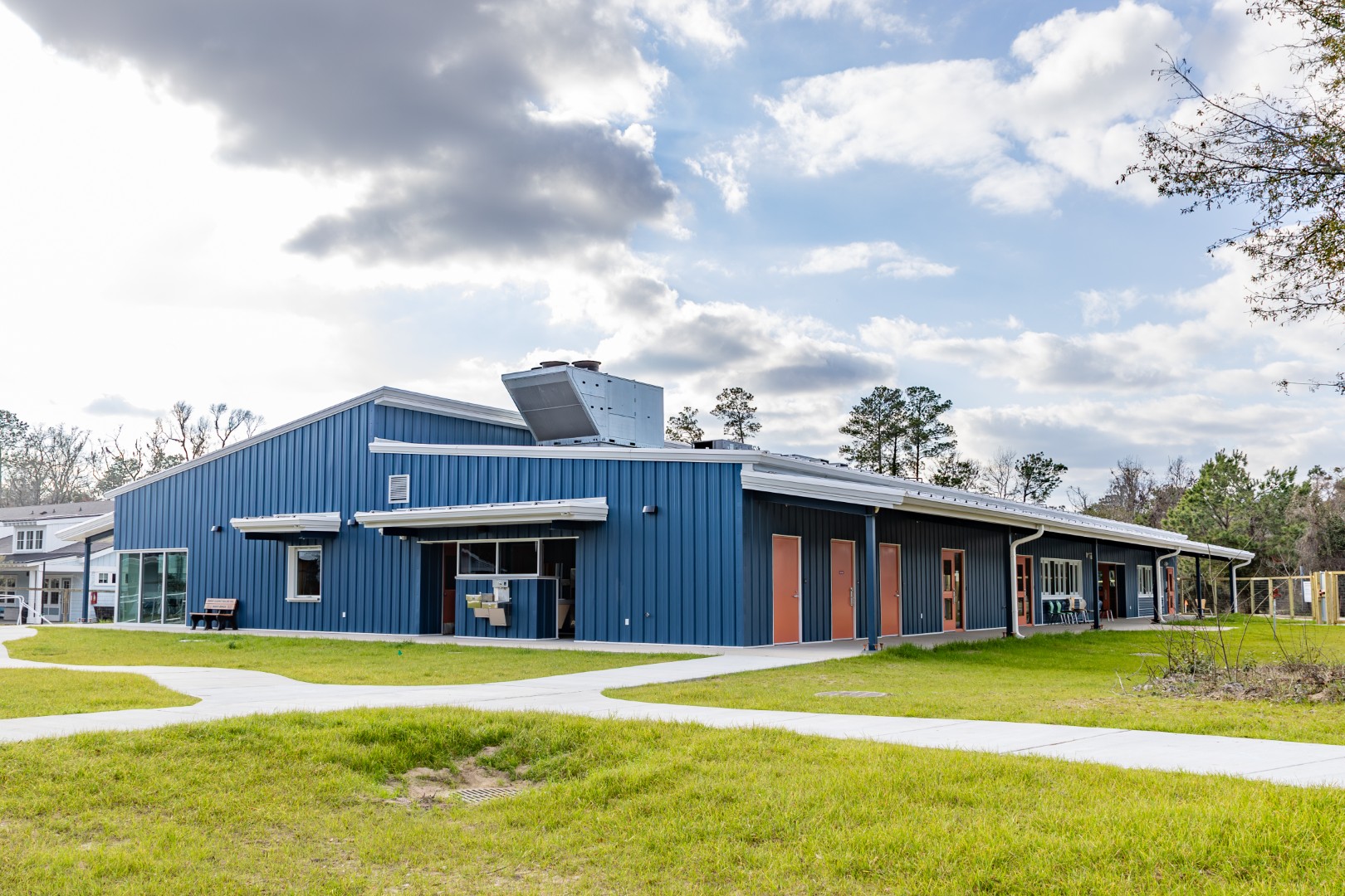 A single-story building with blue metal siding and large windows, surrounded by green lawns and trees under a partly cloudy sky. Concrete walkways curve through the grass in the foreground.