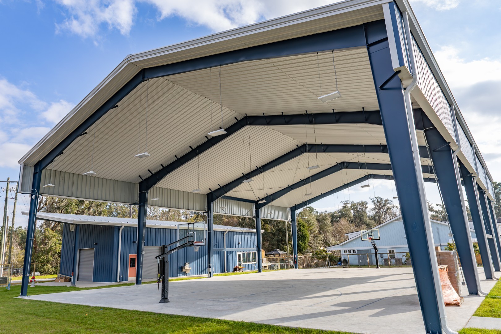 A large, open-sided covered basketball court with metal beams and a high roof stands on a concrete surface, with two basketball hoops and blue buildings in the background under a partly cloudy sky.