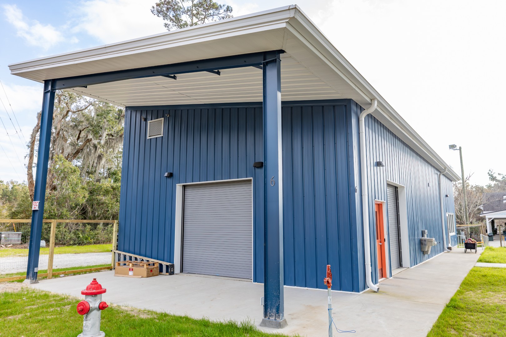 A blue metal building with a covered awning, gray roll-up door, and orange side door sits on a concrete slab. A red fire hydrant and grassy area are in the foreground. Trees and a fence are in the background.