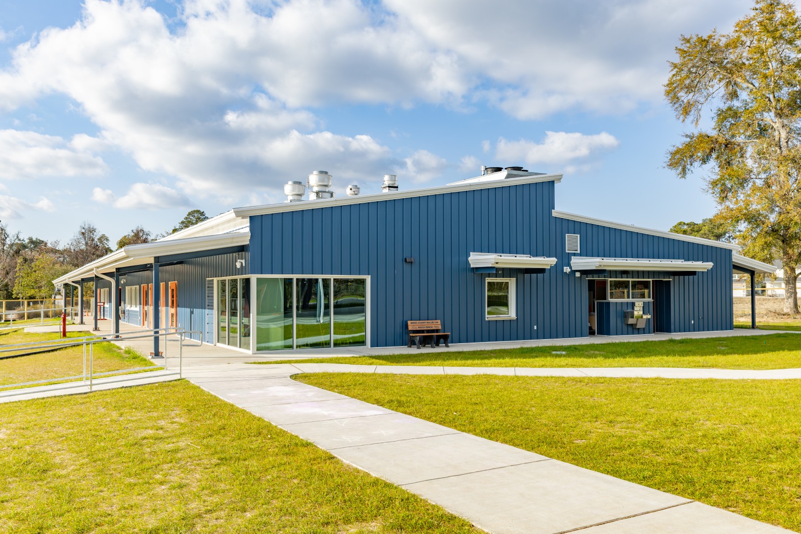 A blue building with large windows and multiple entrances sits on a grassy area under a partly cloudy sky, with sidewalks leading to the doors and a bench near the entrance.