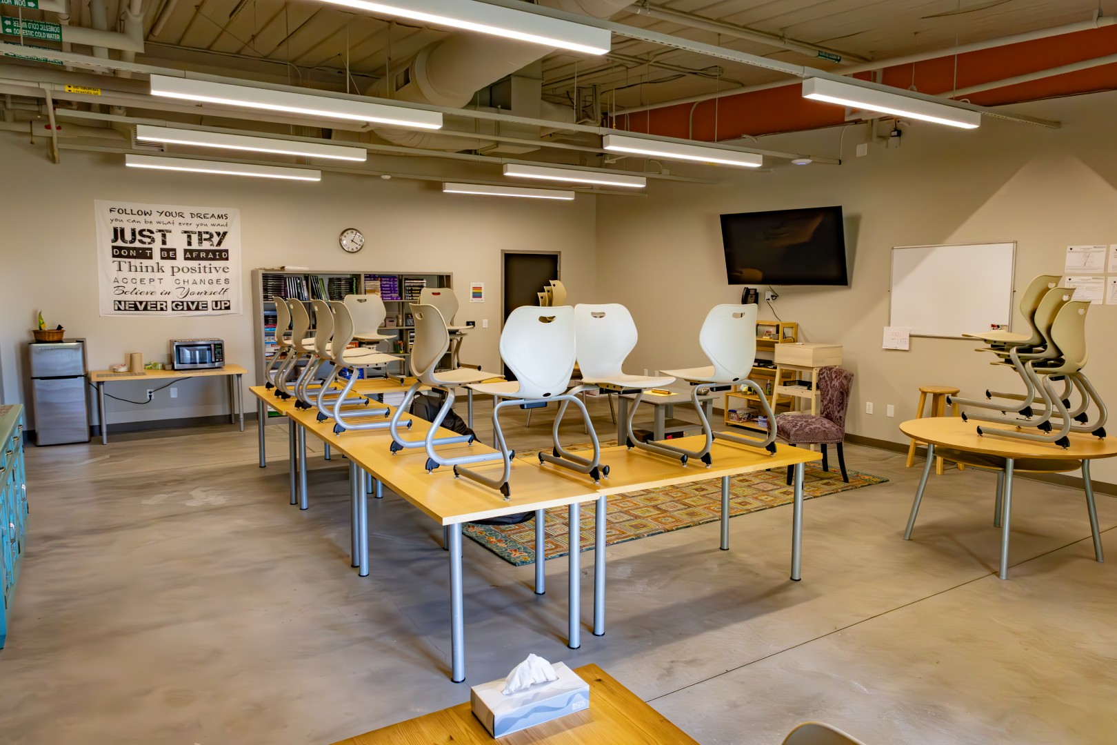 A classroom with empty chairs stacked upside-down on tables, motivational posters on the wall, a flat-screen TV, whiteboard, bookshelf, and bright overhead lighting. The room appears clean and organized.