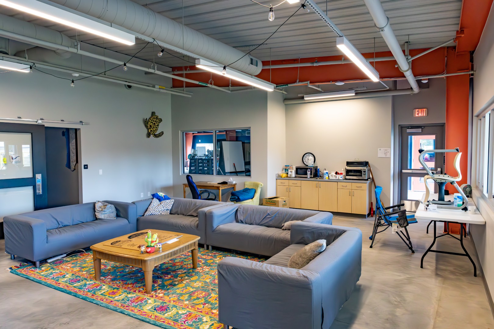 Lounge area with gray sofas around a wooden coffee table on a colorful rug, office chairs, cabinets, and various office equipment in a room with exposed ceilings and large windows.