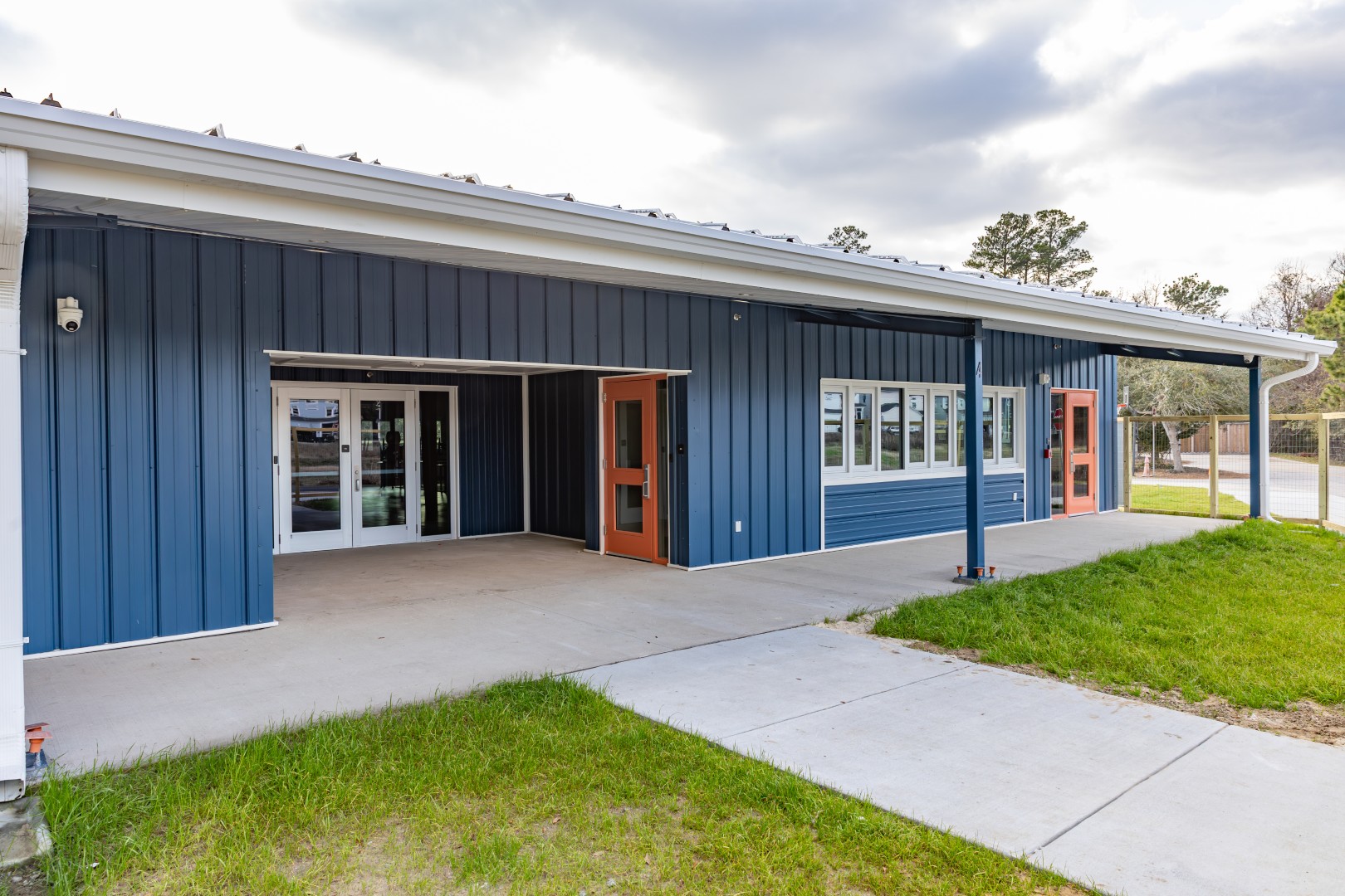 A single-story building with blue siding, large windows, and orange doors. The building has a covered walkway, concrete patio, and is surrounded by grass and a partly cloudy sky.