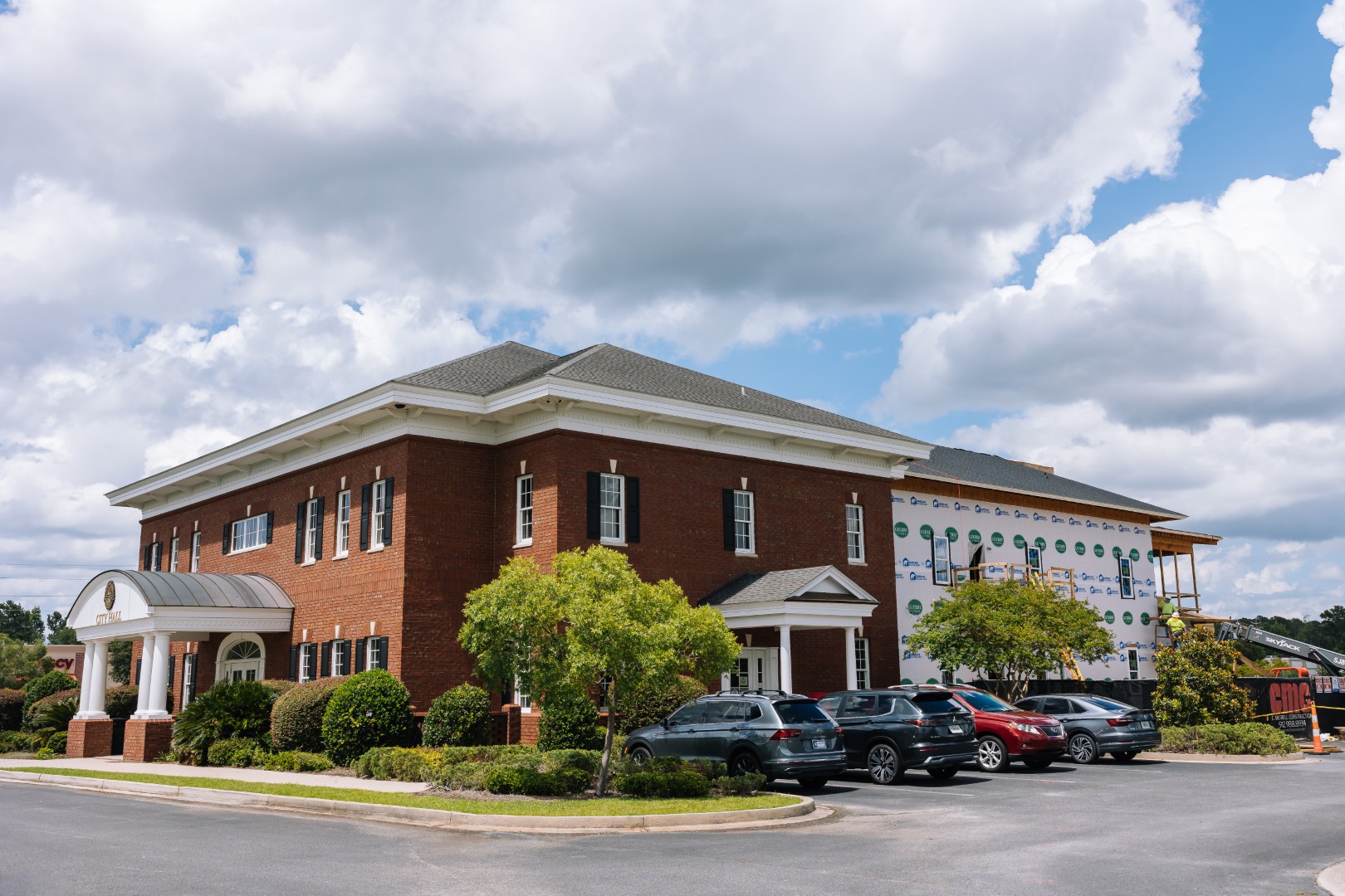 A large red-brick office building with several windows; part of the building is under construction, covered with house wrap. Multiple cars are parked in front, and the sky above is partly cloudy.