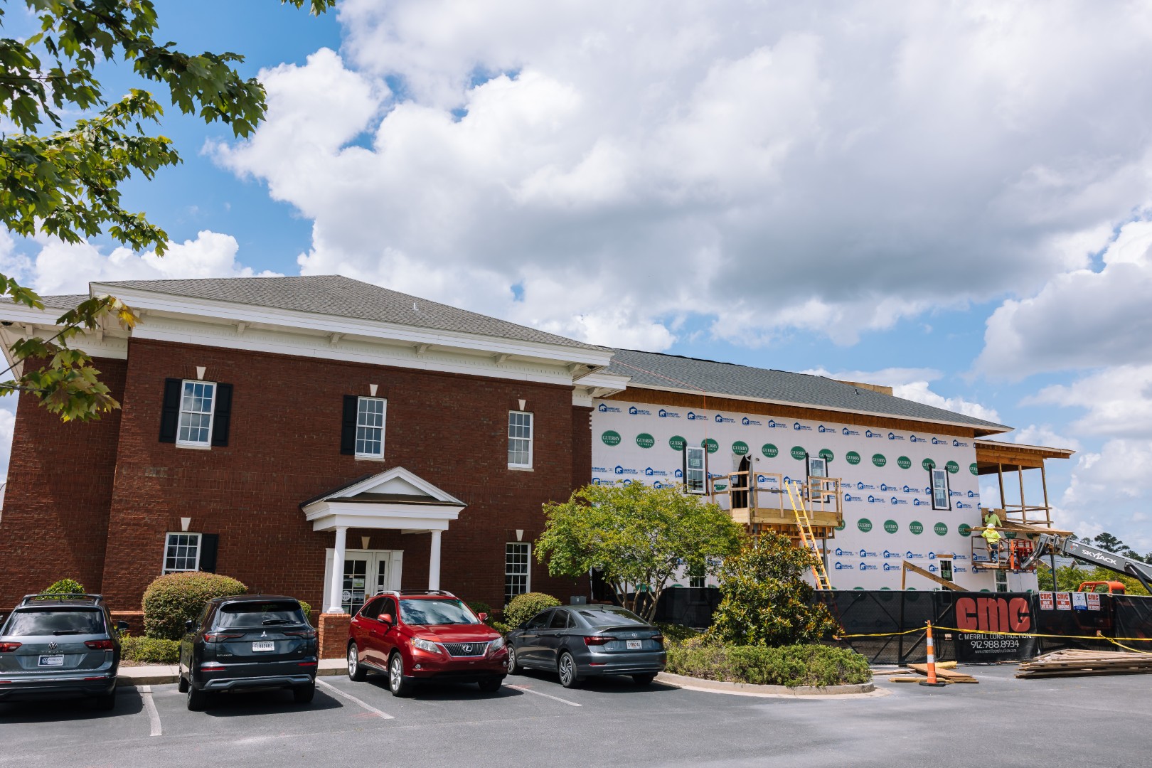 A red brick building under construction, with part of its exterior covered in house wrap. Several cars are parked in front, and construction equipment and scaffolding are visible on the right side.